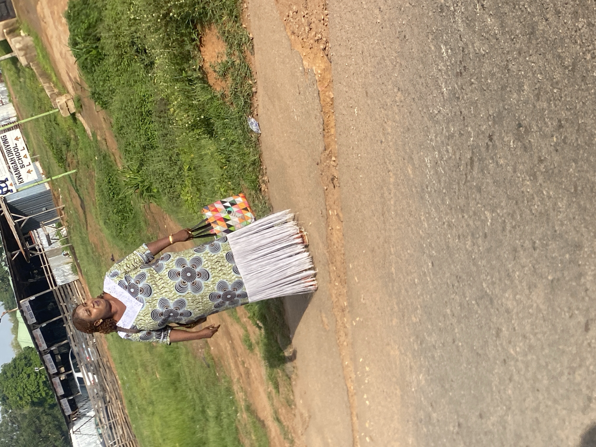 A middle-aged Black woman with braided hair walks along a dusty path next to a road. She is wearing a long, patterned dress with a white, v-shaped neckline and a fringed hem. In her right hand, she carries a multi-colored tote bag. The background shows a grassy area with some scattered white flowers, a sign for "KWANGAH DRIVING SCHOOL," and several structures, possibly part of a market or workshop, with corrugated metal roofing and wooden fencing. The overall scene appears to be in an outdoor, possibly semi-rural, setting within the city of Jos, Nigeria, suggested by the presence of local businesses and vernacular architecture.
