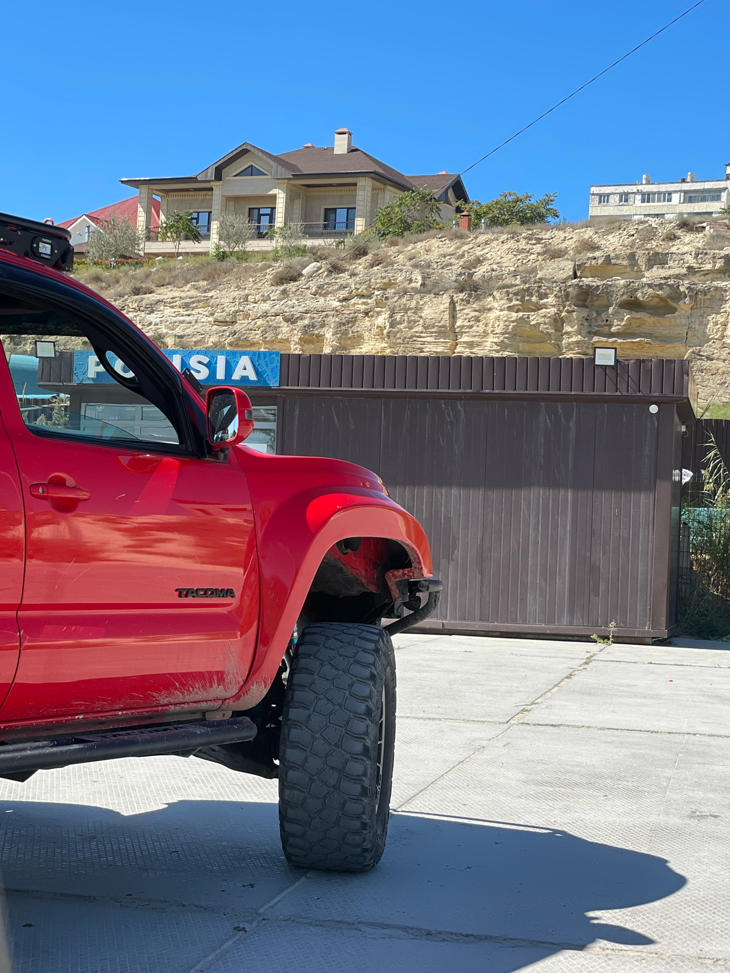 A red Toyota Tacoma pickup truck is parked on a concrete surface in front of a large, brown corrugated metal fence. The truck is shown from the side, with its front quarter and driver's side door visible. The word "TACOMA" is visible in black lettering on the truck's door. Behind the fence, a steep, rocky embankment rises up, topped with several buildings. The sky is clear and blue.