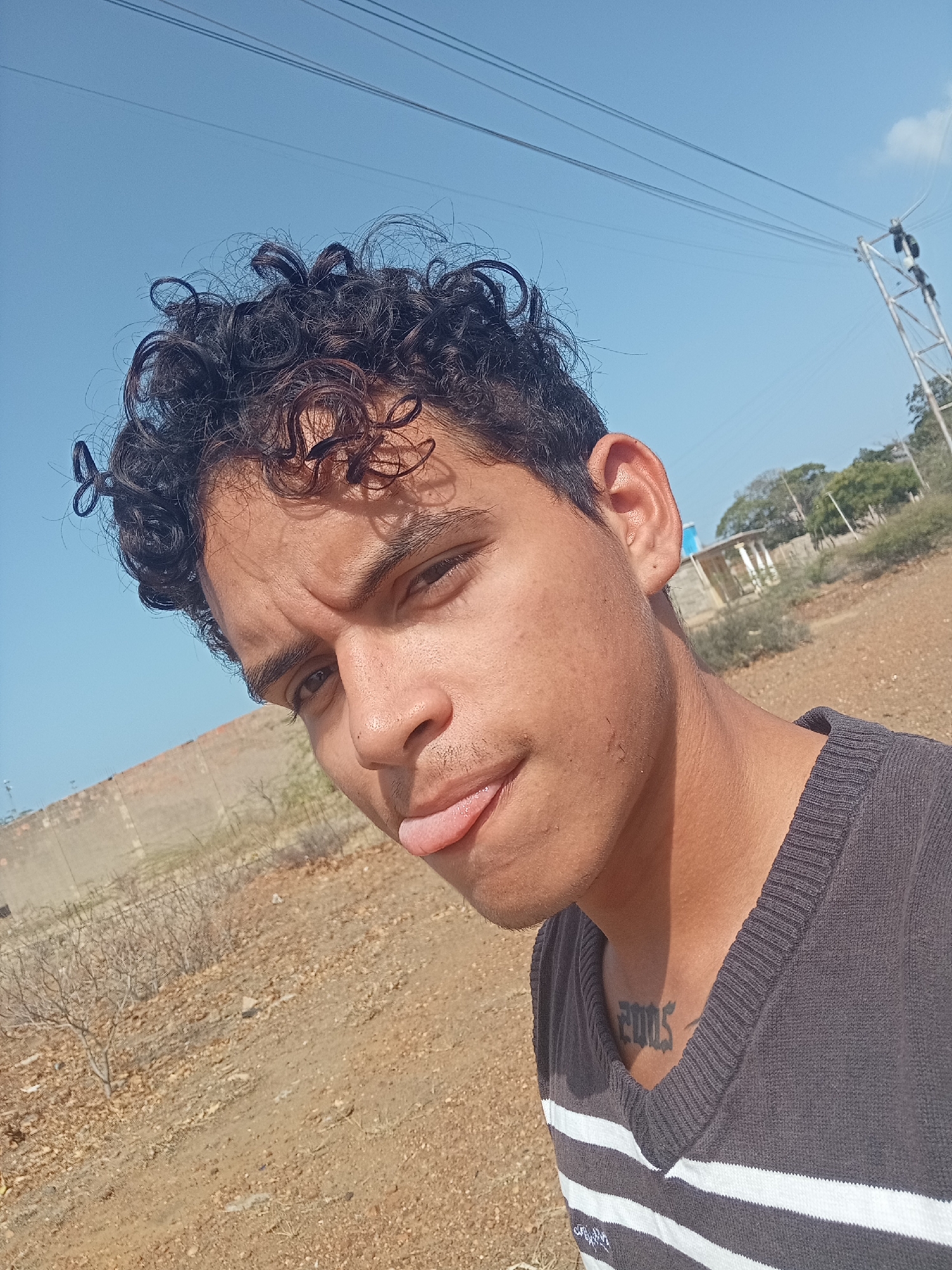 The image is a close-up, angled selfie of a young man, taken outdoors in Altagracia, Venezuela, under bright daylight. The subject has dark, curly hair and dark eyes, looking directly at the camera with a playful expression, his tongue slightly protruding. His eyebrows are lightly furrowed, possibly due to the sun or his expression. He wears a dark gray V-neck sweater featuring white horizontal stripes on the lower half. A tattoo with the numbers "2005" is visible on his upper right chest.
The background shows a vast, clear blue sky with a hint of a small cloud near a distant utility pole. The ground beneath is dry, dusty, and light brown, characteristic of a semi-arid environment, with sparse, dry vegetation including some bare bushes and small trees. In the distance, a low, light-colored wall or structure is visible on the left, and power lines stretch across the sky towards a metal utility pole on the right. The scene suggests a sunny, clear day in a rural or unpaved outdoor setting.