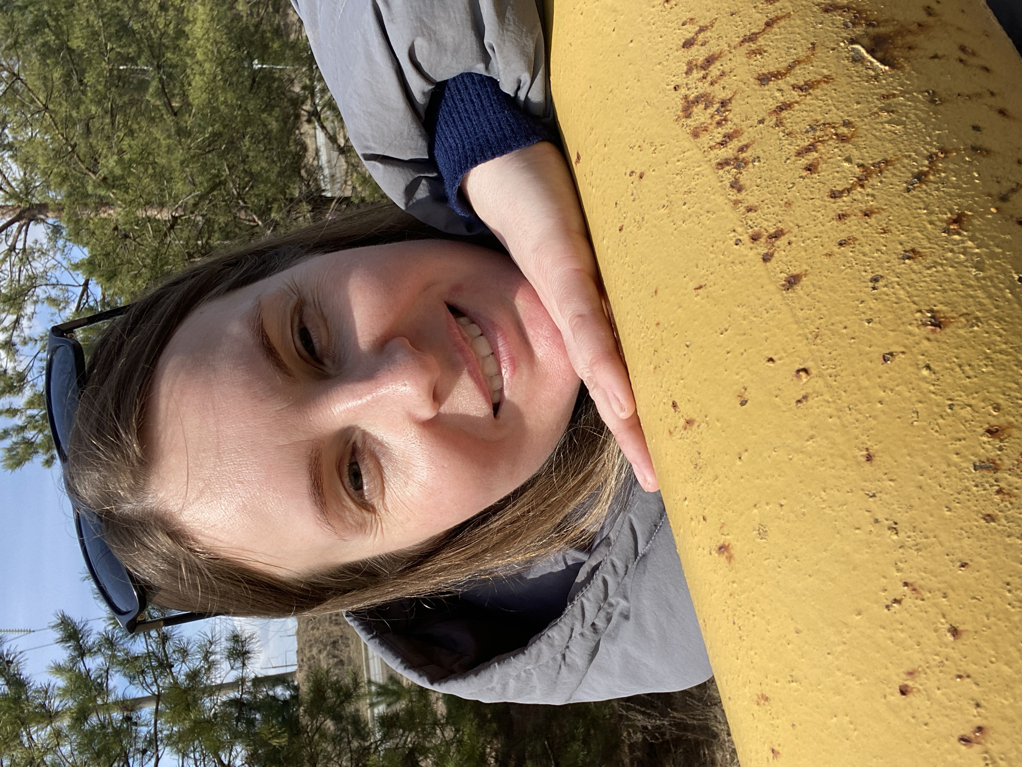 A fair-skinned woman with light brown hair is seen in a close-up, vertically oriented shot. She is wearing a grey jacket with a blue knit sleeve visible at the cuff. She is smiling and resting her face and hand against a rusty yellow metal post. Sunglasses are tucked into her hair near her forehead. The background shows trees and a blue sky with some white clouds. The location is Noviy, Russia.