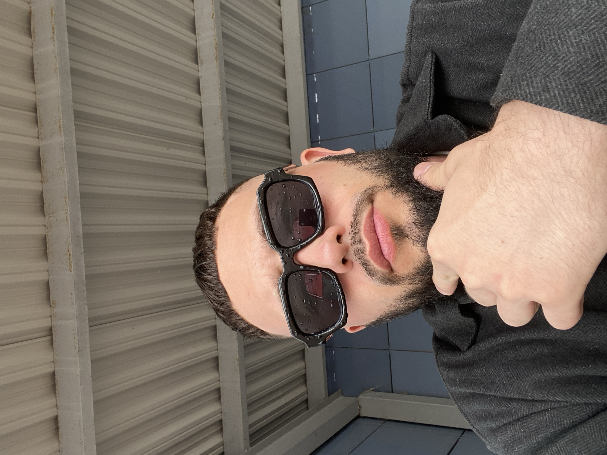 The image shows a close-up, low-angle, portrait-style shot of a man wearing black sunglasses and a dark gray coat. The man has a beard and is touching his chin with his right hand. His mouth is slightly open, revealing his teeth and pink lips. The sunglasses have water droplets on them, suggesting it might be raining or has recently rained.
The background consists of corrugated metal siding and dark blue tiled walls. The overall lighting suggests it is daytime, possibly overcast due to the rain. The setting appears to be outdoors, perhaps under a covered walkway or shelter, given the siding and the tiled wall.
There is no visible text in the image. The man's expression is neutral, and the focus is on his face and hand. The image has a slightly grainy texture. The crops show more detail of the man's features, the texture of his clothing, and the background elements. The location context of Chaikovsky, Russia, does not provide any specific visual cues within the image itself.