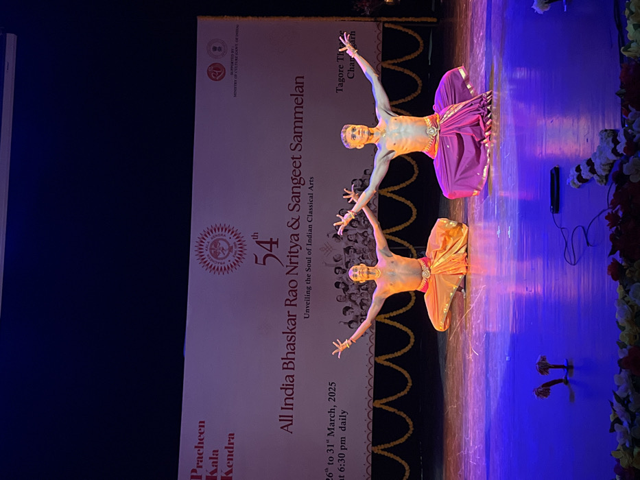 Two male dancers are performing on a stage at the Tagore Theatre in Chandigarh, India, at the 54th All India Bhaskar Rao Nritya & Sangeet Sammelan. The event is being held from 26th to 31st March, 2025, and is supported by the Ministry of Culture, Government of India. Both dancers are wearing traditional Indian attire, with one in an orange dhoti and the other in a purple dhoti. They are both in a seated position, with their arms outstretched in a dance pose. The stage is lit with blue lights and there are garlands and a black cable on the floor in front of the dancers. The backdrop is a white banner with the event details and logo. The coordinates provided are for the Tagore Theatre in Chandigarh, India.