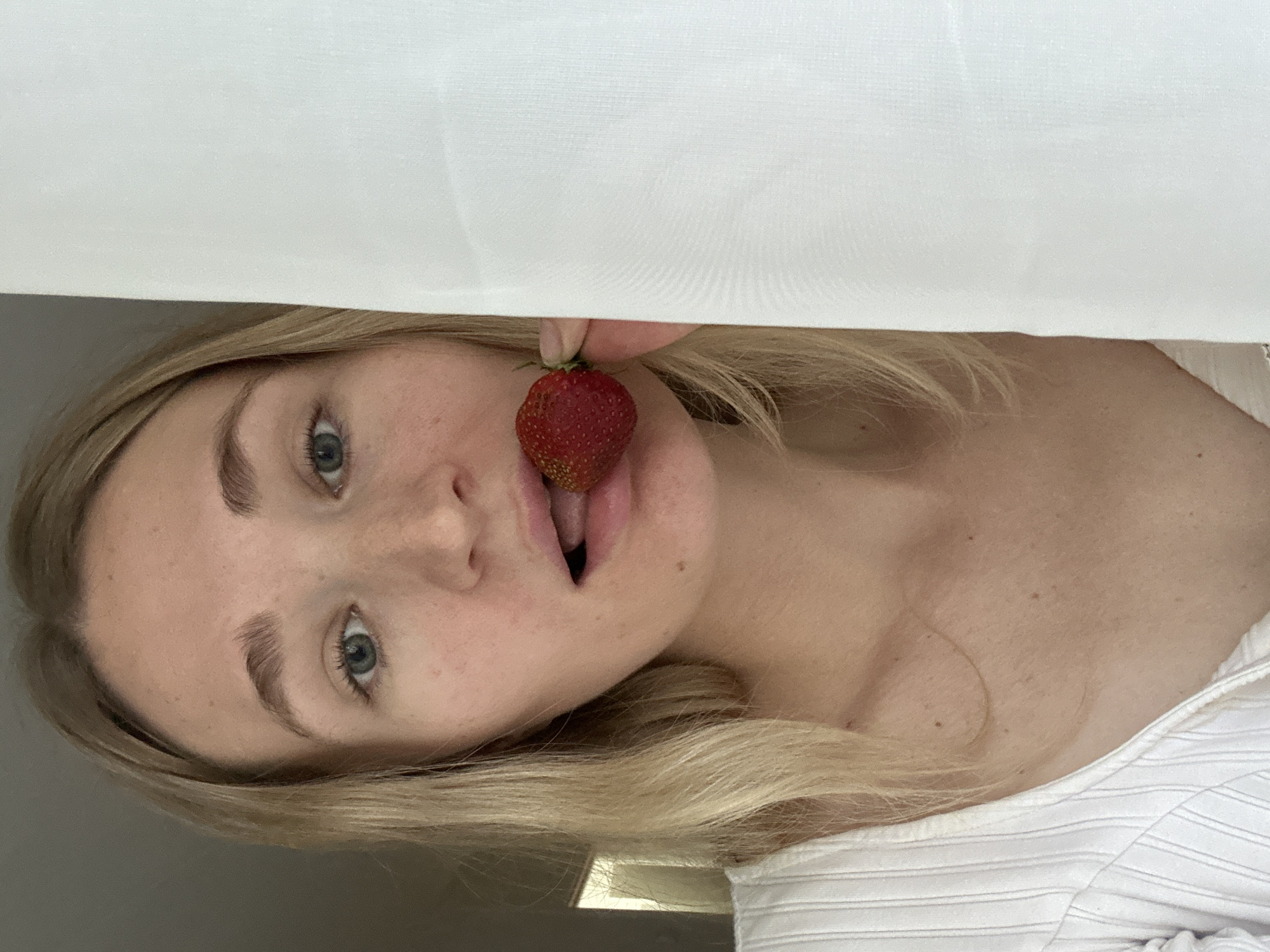 A blonde woman, possibly in her early twenties, is positioned in the foreground, looking directly at the camera. She has fair skin with some freckles, light eyebrows, and blue eyes. Her mouth is slightly open, and she is holding a single ripe strawberry to her lips with her right hand, her index finger and thumb delicately grasping its stem. A corner of a white, sheer curtain is visible to her right, partially obscuring her face and shoulder. The background to her left is dark and indistinct, suggesting an indoor setting, possibly near a window or a doorway. She is wearing a white, ribbed garment that appears to be a top or dress. There are no discernible city landmarks or environmental features to indicate a specific location in Chaikovsky, Russia.