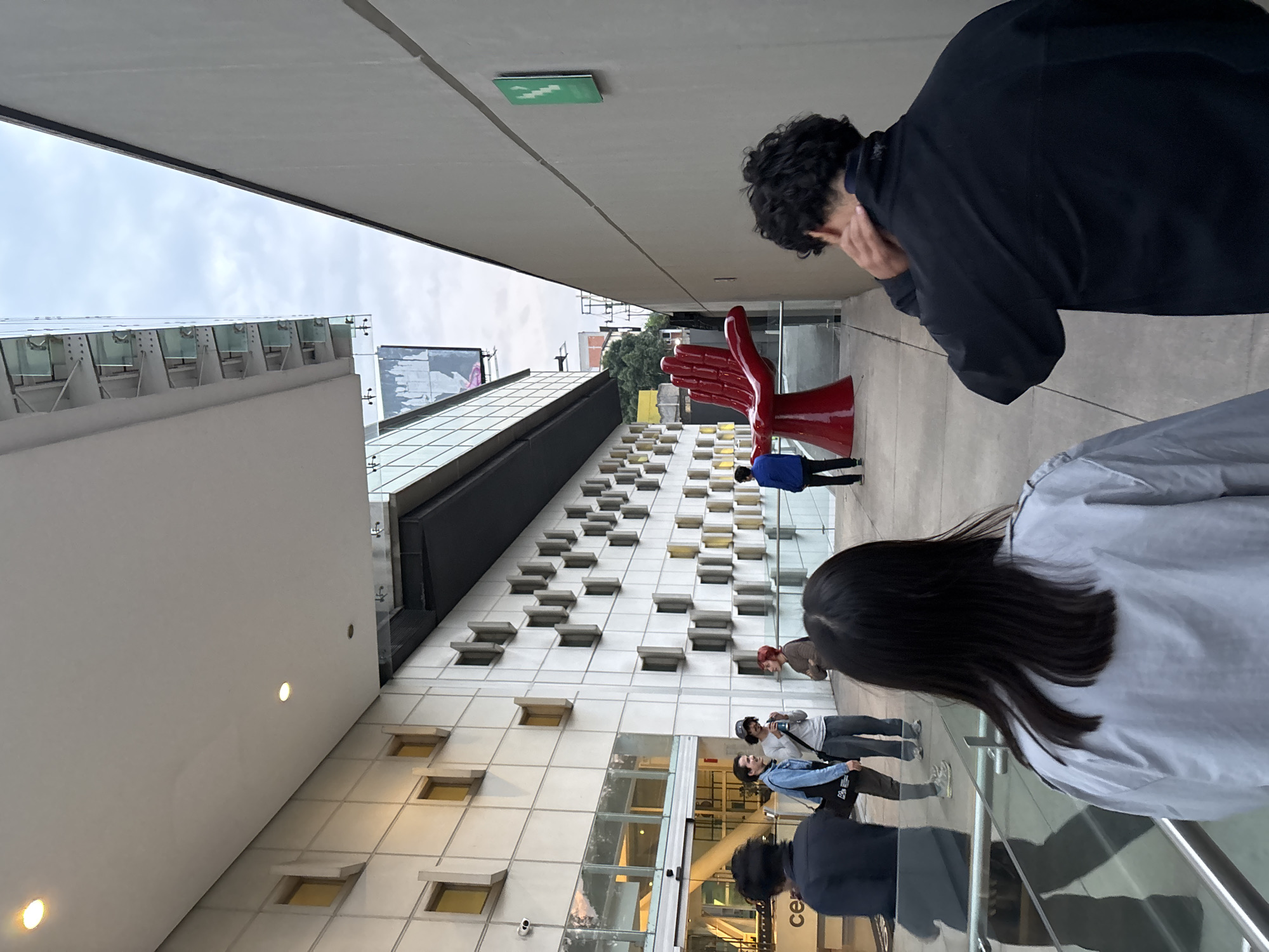 The image, taken in Mexico City, Mexico, shows a pedestrian walkway between two buildings under an overcast sky. A red, stylized sculpture of two hands is positioned along the walkway. Several people are present: one in a blue jacket, two taking photos near the entrance of a building, and two more partially visible in the foreground. The architecture is modern, with a white building featuring rows of small windows and glass paneling. A large advertisement is visible atop one of the buildings.