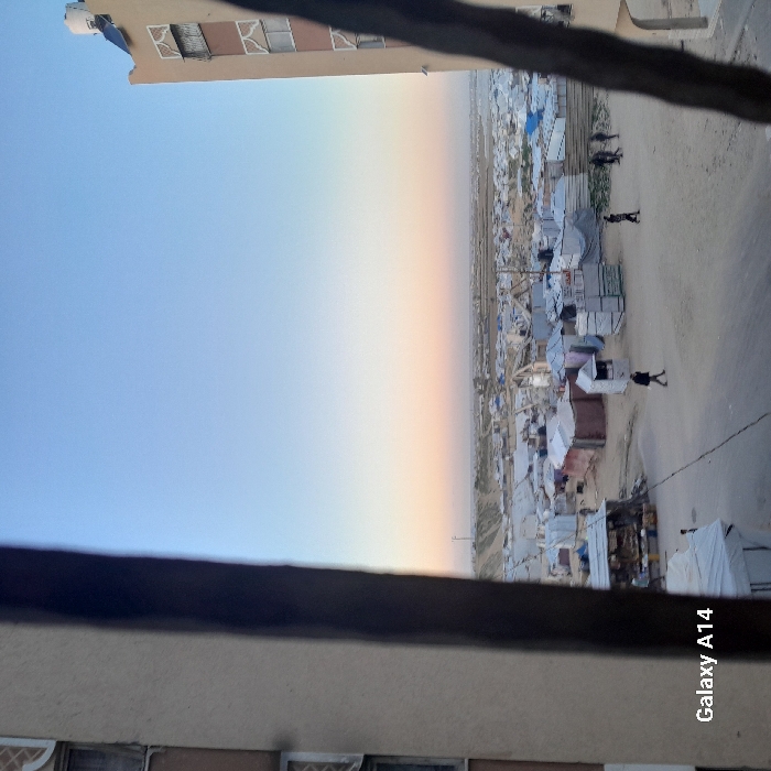 The image captures an expansive outdoor scene, viewed from an elevated perspective, likely from a window or balcony. The photograph is oriented vertically but depicts a landscape that appears to have been taken horizontally and then rotated. When viewed upright (rotated 90 degrees clockwise), a dark horizontal bar, resembling a railing or window frame, runs across the bottom section of the image, partially obscuring the lower view. Below this bar, the light brown/beige exterior wall of a building is visible, featuring several windows or vents.
To the left side of the frame, a portion of a light brown building rises, displaying multiple windows or openings, some fitted with shutters or blinds. On its roof, a metallic object, possibly a solar water heater or an antenna, can be seen.
The dominant feature from the mid-ground extending to the distant horizon on the right is a sprawling settlement or camp. It is densely packed with numerous small, irregularly shaped structures, appearing to be tents or makeshift shelters, primarily in shades of white, grey, and light blue. Some darker, more solid-looking buildings are also interspersed within the camp. Unpaved dirt paths or roads wind through the settlement. Several small, dark figures, presumably people, are visible walking or standing within the camp area, indicating human presence and activity.
The sky occupies the upper portion of the image, displaying a gentle gradient of colors. It transitions from a clear, light blue at the top to a soft peach, orange, and yellowish hue closer to the distant, flat horizon line. This coloration suggests either a sunrise or a sunset. The horizon stretches far into the distance, indicating a vast, open landscape, possibly a plain or desert.
In the bottom right corner of the re-oriented image, the white text "Galaxy A14" is visible, indicating the device used to capture the photograph.