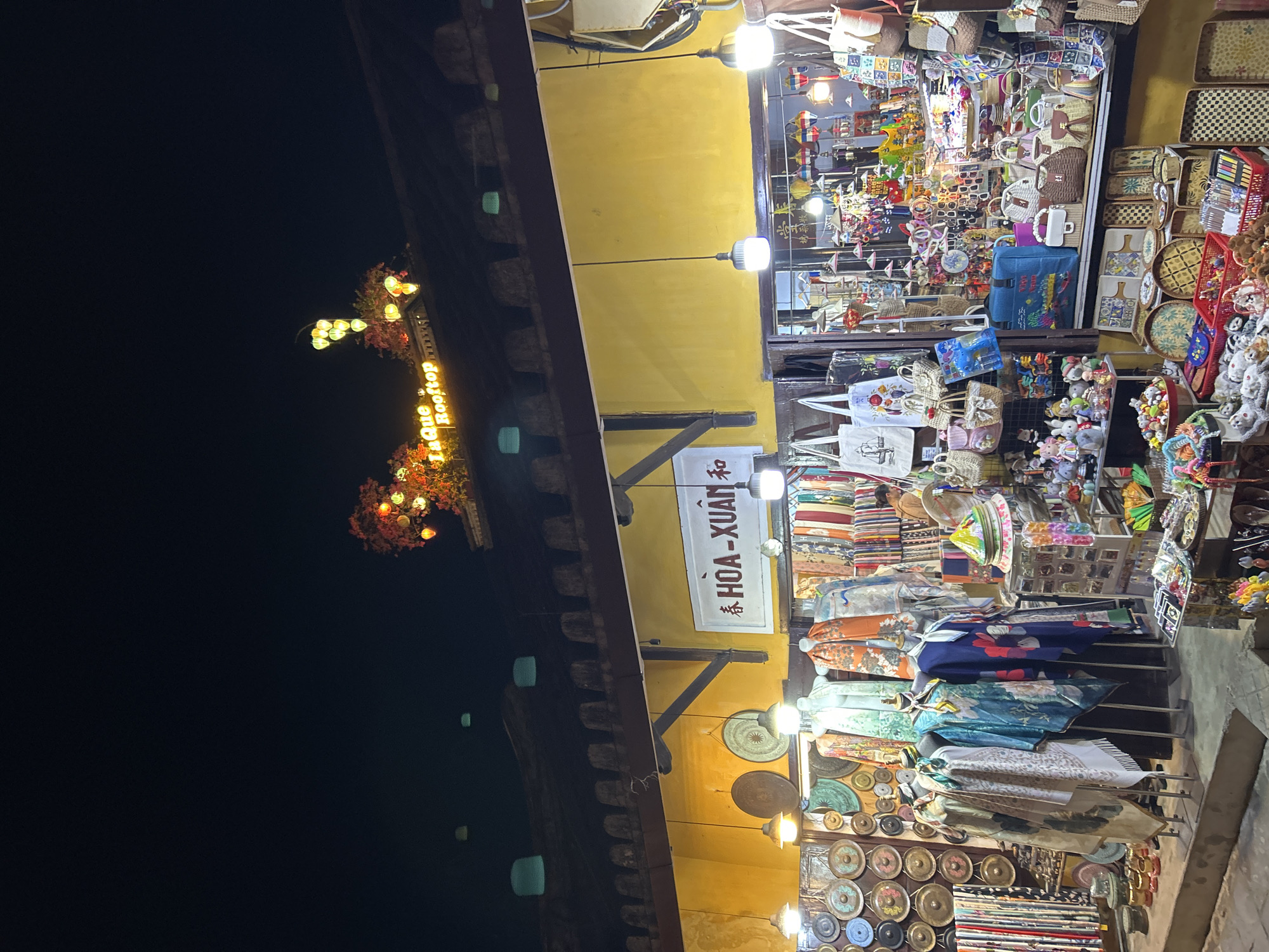 This is an exterior nighttime photograph of a shop in Hội An, Vietnam, which is likely a souvenir or textile shop. The shop features a yellow facade with a dark brown roofline adorned with decorative elements and lights. A sign above the entrance reads "HÒA-XUÂN" in white lettering on a dark background. The shop is packed with various items for sale, including textiles like scarves and clothing draped over mannequins, woven baskets and bags, decorative plates, and an assortment of small trinkets and stuffed animals. Several string lights and decorative lanterns are visible, illuminating the merchandise and the storefront. The overall impression is one of a vibrant and busy night market scene.