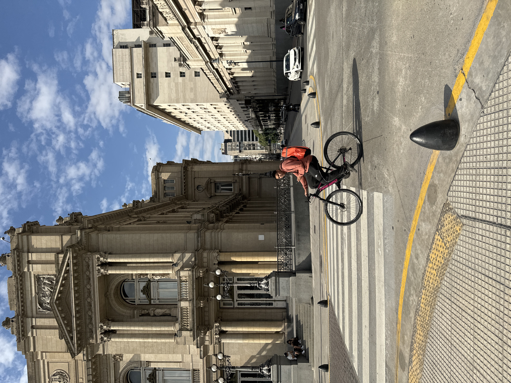 This vertical image captures a bustling street scene in Buenos Aires, Argentina, under a bright, partly cloudy blue sky. The strong sunlight casts distinct shadows, suggesting it's either mid-morning or afternoon.
The primary subject is a person on a black bicycle, standing on a wide, striped pedestrian crossing in the center-right of the frame. They are wearing a peach or light orange hoodie and a prominent bright orange backpack. The bicycle frame clearly displays the brand name "FIREBIRD". To the far left, two other individuals are seated on the steps of a grand, classical-style building that occupies much of the left side of the image.
This imposing building features elaborate architectural details, including multiple columns, decorative statues, and a prominent pediment above an arched entrance. Ornate streetlights are visible on its facade. The street itself is concrete, bordered by a bright yellow painted line and a textured sidewalk with tactile paving. Several black, conical bollards line the curb.
Further down the street, in the middle ground, another large, multi-story building with a similar classical architectural style is visible. Faint text, possibly "BANCO DE LA NACION," can be discerned on its upper facade. Two cars, one white and one dark, are parked along the street. The overall setting conveys an active urban environment rich with historical architecture.