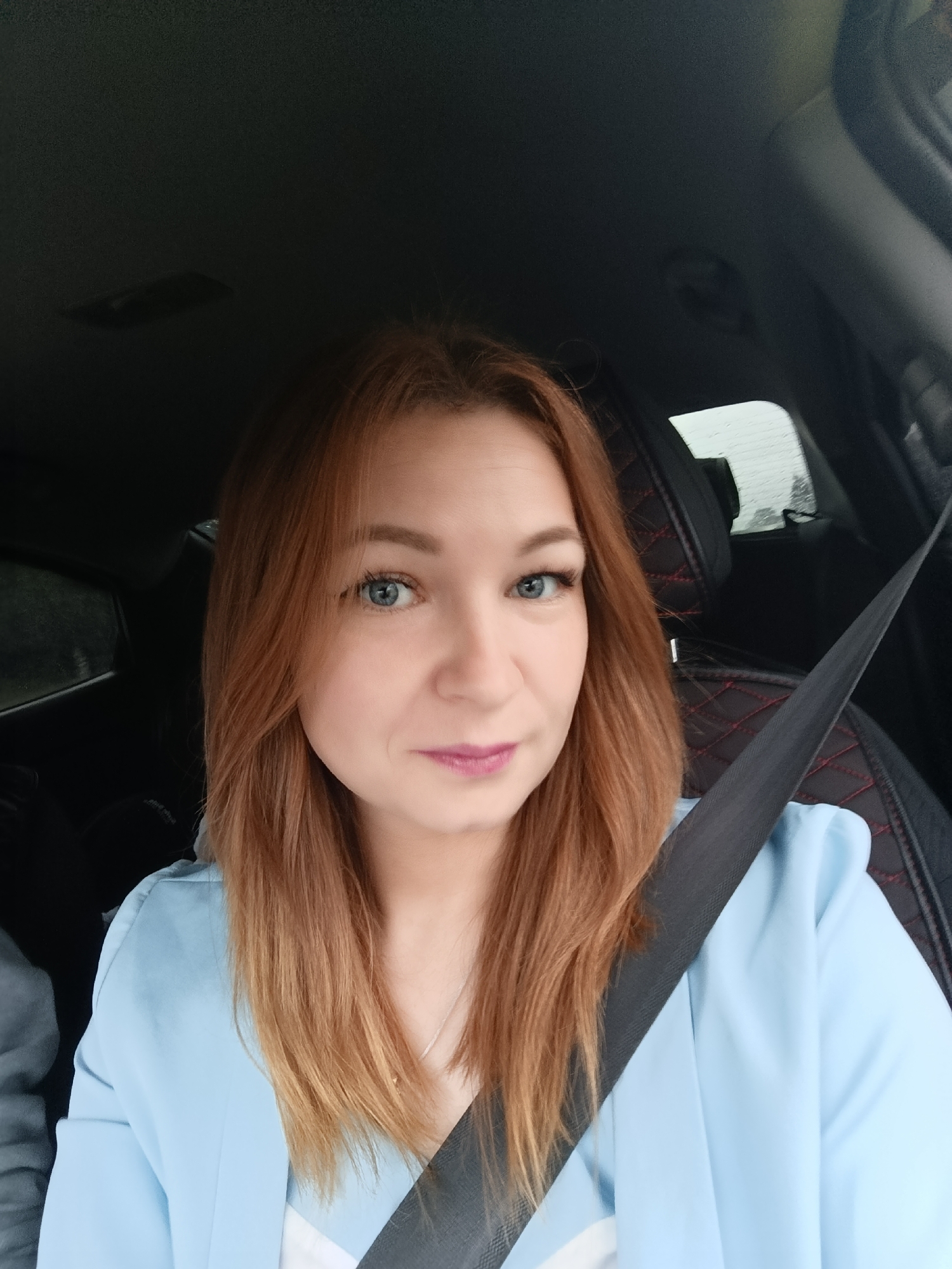 A medium shot captures a young woman with auburn hair sitting in the driver's seat of a car, looking directly at the camera. She is wearing a light blue blazer and a black seatbelt. The car's interior is visible, including the black leather seats with red stitching and the car's roof lining. Rain streaks are visible on the car window in the background. The image appears to have been taken outdoors, possibly on a cloudy day, in the city of Chaikovsky, Russia, given the context.