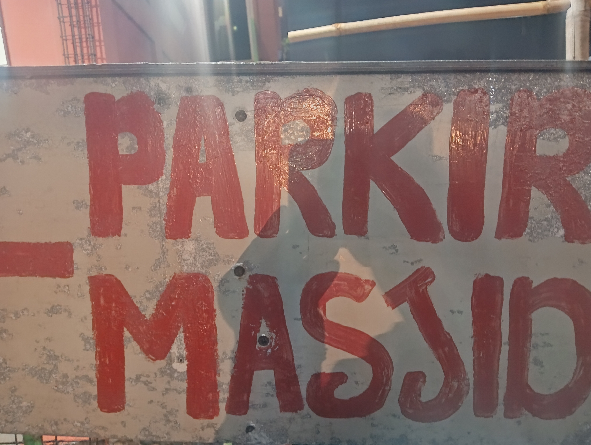 The image shows a weathered sign with red painted lettering indicating "PARKIR MASJID", which translates to "MOSQUE PARKING" in English. The sign is mounted outdoors, suggesting it is in a public area. The background is blurred but appears to be a somewhat urban or semi-urban environment with some construction elements and greenery. The sign itself shows signs of age and exposure to the elements, with some chipping and wear on the painted surface. There are no people visible in the image. The location is specified as Cisarua, Indonesia, which is a popular tourist destination in West Java, known for its natural beauty and as a gateway to the Puncak Pass.