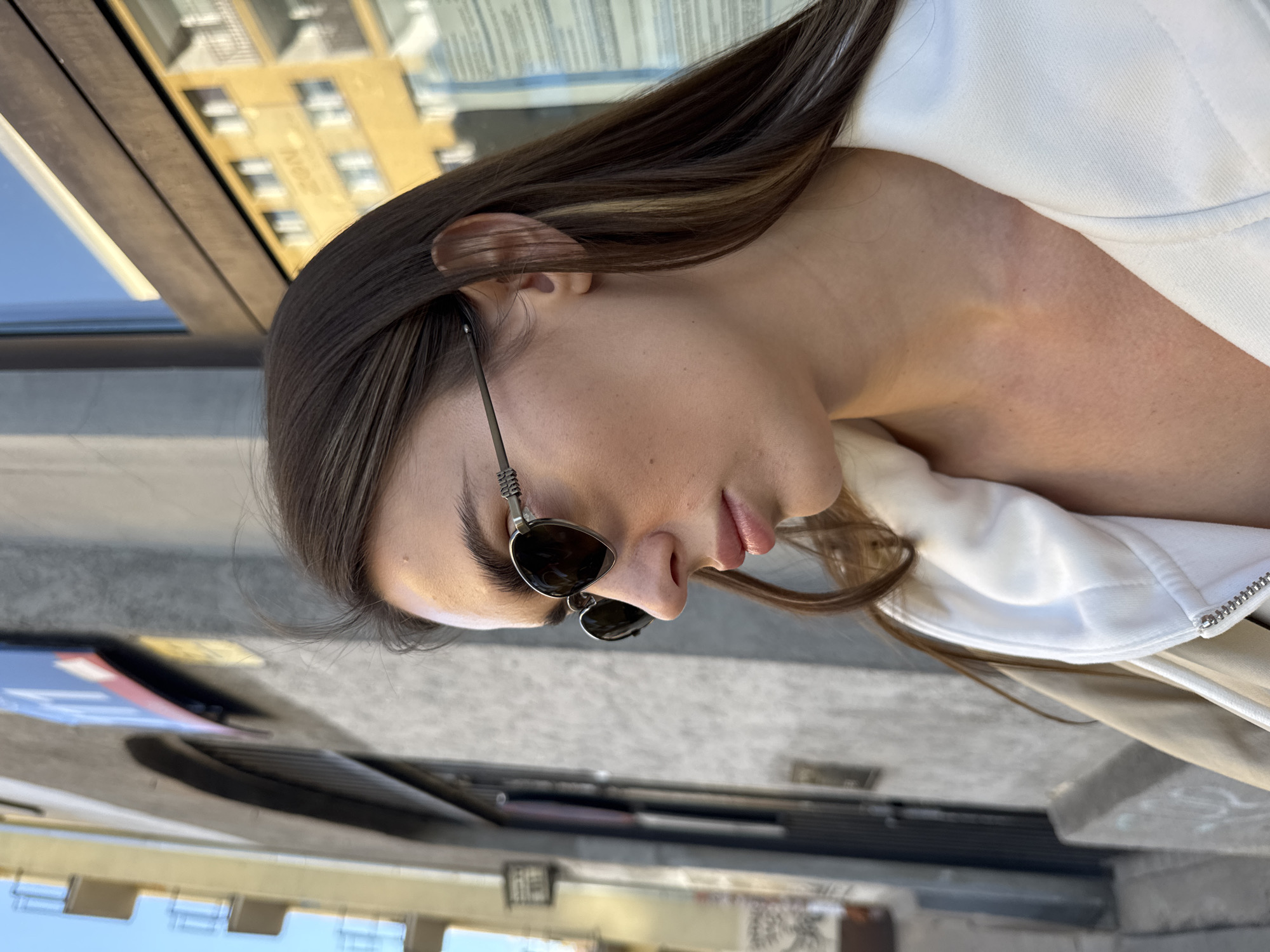 A woman wearing sunglasses is seated at an outdoor table, possibly at a cafe or restaurant. She is wearing a white jacket. The setting appears to be in a city; the reflection in a nearby window shows buildings. Given the context of Warsaw, Poland, the buildings may reflect the local architecture. The focus of the shot is on the woman's face and upper body. The table in front of her appears to be a dark color.