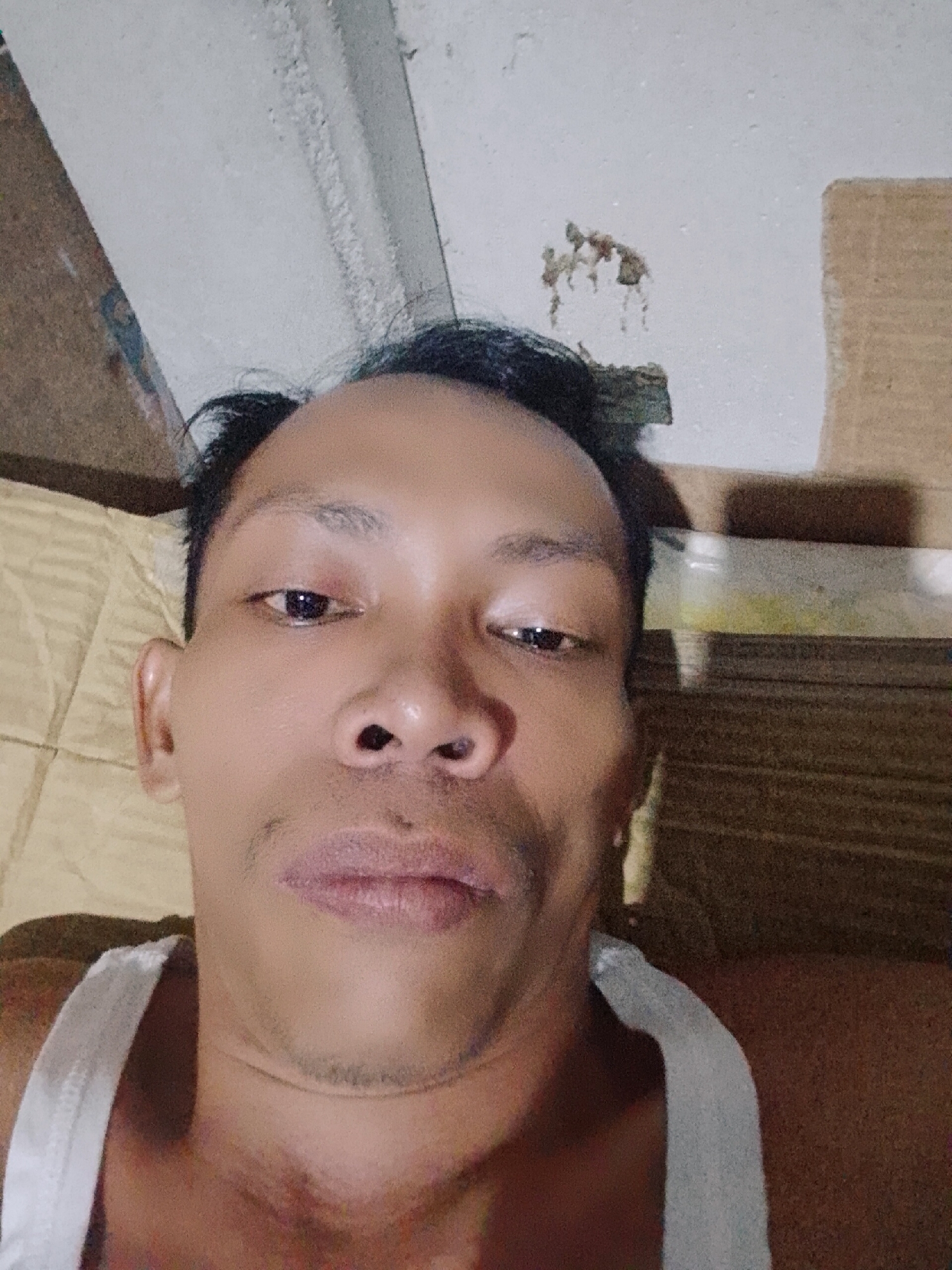 A selfie of a person with dark hair and brown skin wearing a white tank top. The person is lying down on a yellow surface. Behind the person are sections of a cardboard box and a white painted ceiling. The location is possibly Bukit Ketok, Indonesia.