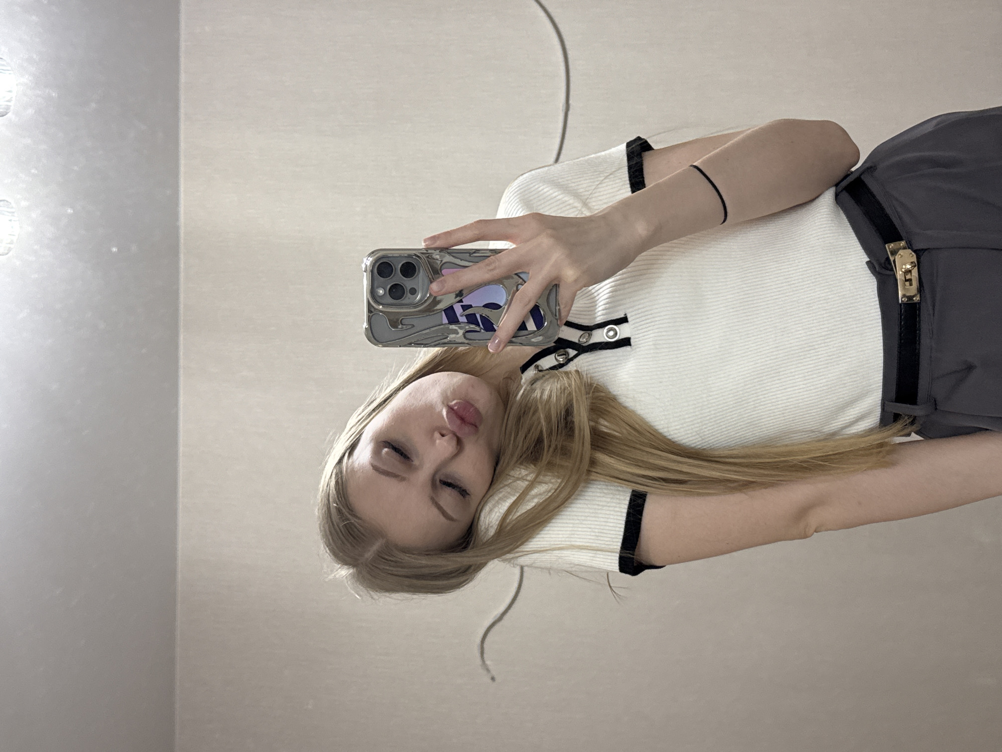 A young woman with long blonde hair is taking a selfie with a smartphone with a silver-colored decorative case. She is wearing a white knit top with black trim and dark-colored pants or a skirt with a belt. The background appears to be a plain wall, possibly inside a building. The image is slightly rotated, and there is a thin, curved line visible near the top of the frame. There is no information in the picture to indicate this image was taken in the city of Perm, Russia.