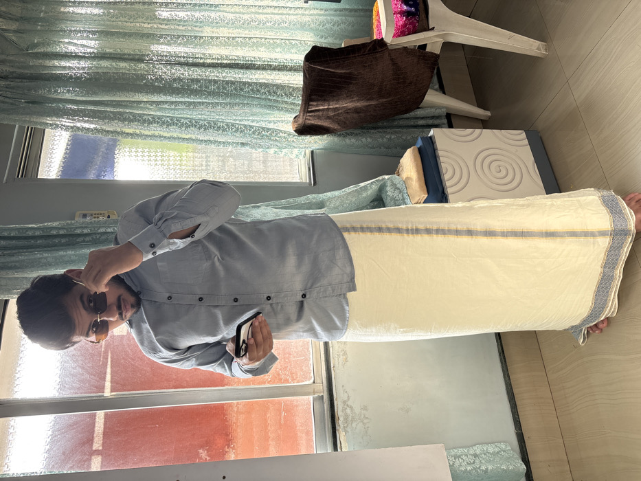 A single male subject stands indoors, centered in the frame. He is light-skinned, with dark hair and a beard. He wears a light blue long-sleeved collared shirt and a cream-colored traditional lower garment (dhoti/lungi) with a blue and grey striped border and a vertical yellow and grey stripe. He is barefoot. The man wears rectangular dark-tinted sunglasses, which he is pulling down with his left hand to peer over them while looking directly at the camera. He holds a white smartphone in his right hand. The background includes two windows, one showing a reddish-orange exterior hue and the other partially covered by sheer light blue patterned curtains, revealing bright daylight. To the right, a white plastic chair is draped with a brown fabric and holds a colorful patterned cloth. Adjacent is a white decorative stand with concentric circle patterns, topped with folded dark blue and beige textiles. A white panel is partially visible on the far left. The floor is light-colored tiled. This indoor scene is located in the city of Howrah, India.