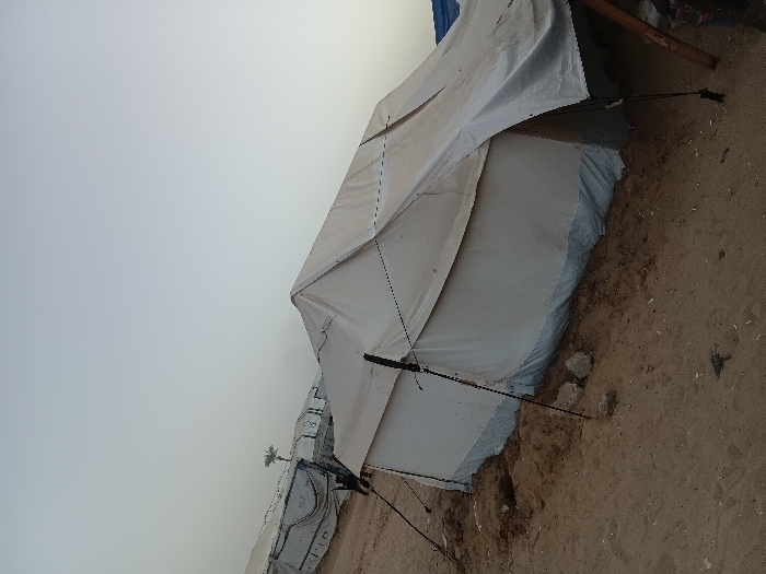 The image captures an outdoor scene featuring several large, light-colored tents under an overcast sky. The primary focus is on a prominent tent in the mid-ground, positioned slightly to the right of the frame. This tent is constructed from a white or very light grey, heavy fabric or tarp material, suggesting a durable, temporary shelter. It has a pitched roof design, with visible black ropes or guy lines extending from its sides to anchor it to the sandy ground. One such rope stretches diagonally from the tent's mid-section towards the lower left of the image. The lower part of the tent appears to be secured or reinforced, possibly by piling sand or dirt against its base, and a bluish tarp-like material can be seen layered along its top edge, suggesting a multi-layered wall or repair.
The ground surrounding the tents is predominantly dry, light brown, sandy earth, with scattered small rocks and debris, indicating a possibly arid or semi-arid environment. To the far right, partially cut off, a section of what appears to be rough wooden beams or supports is visible, hinting at other nearby structures or collected materials.
In the background, further to the left, another similar tent is partially visible, also light-colored and of similar construction, though its full extent is obscured. A small, light-colored object, possibly a small plant or a piece of debris, can be seen clinging to the side of this background tent.
The sky occupies a significant portion of the upper half of the image, presenting a uniform, pale grey-blue color, characteristic of an overcast day or perhaps twilight hours (early morning or late afternoon). There are no distinct clouds, only a flat, diffused light.
The image is captured with a tilted perspective, causing the horizon line to appear diagonally across the frame. There are no people visible, and no specific activity is taking place, although the presence of multiple tents strongly suggests a temporary settlement or camp. No discernible text is visible anywhere in the image.