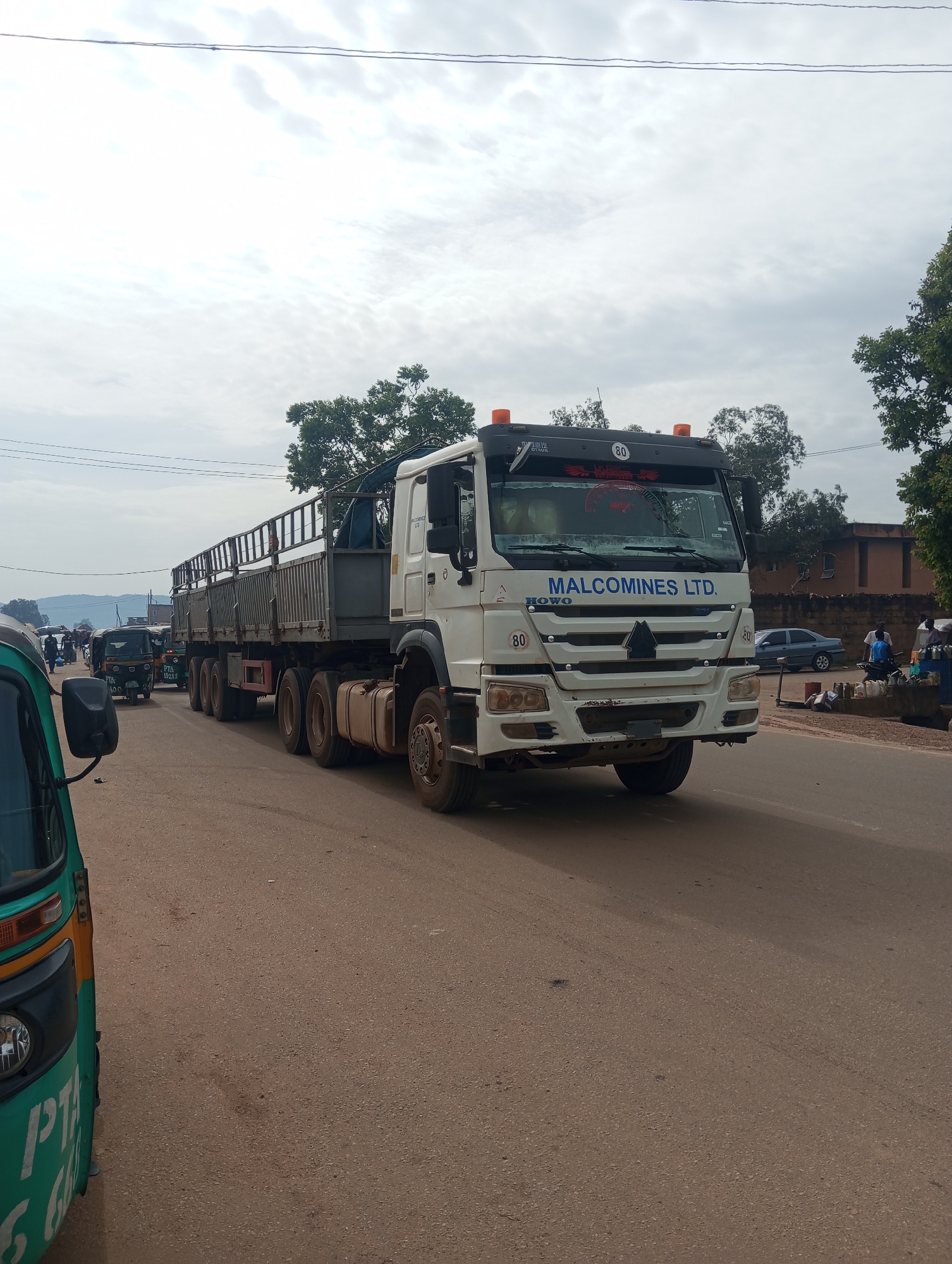 A long, white semi-truck with a trailer is parked on a paved road. The truck has "MALCOMINES LTD." written on the side of the cabin. There are also orange hazard lights on top of the cabin, and a number "80" is visible in multiple places on the truck. The trailer is empty and has a metal railing around the top.
In the background, several other vehicles are visible, including a green and yellow auto-rickshaw in the foreground on the left, and a dark green auto-rickshaw further down the road. There are also people walking and standing on the side of the road. A blue sedan is parked on the right side of the road, near a building that appears to be a shop or stall. The sky is overcast with clouds. The scene appears to be a street in a town or city, likely during daylight hours. The overall impression is one of a busy street with commercial activity. There are trees and buildings visible along the sides of the road.