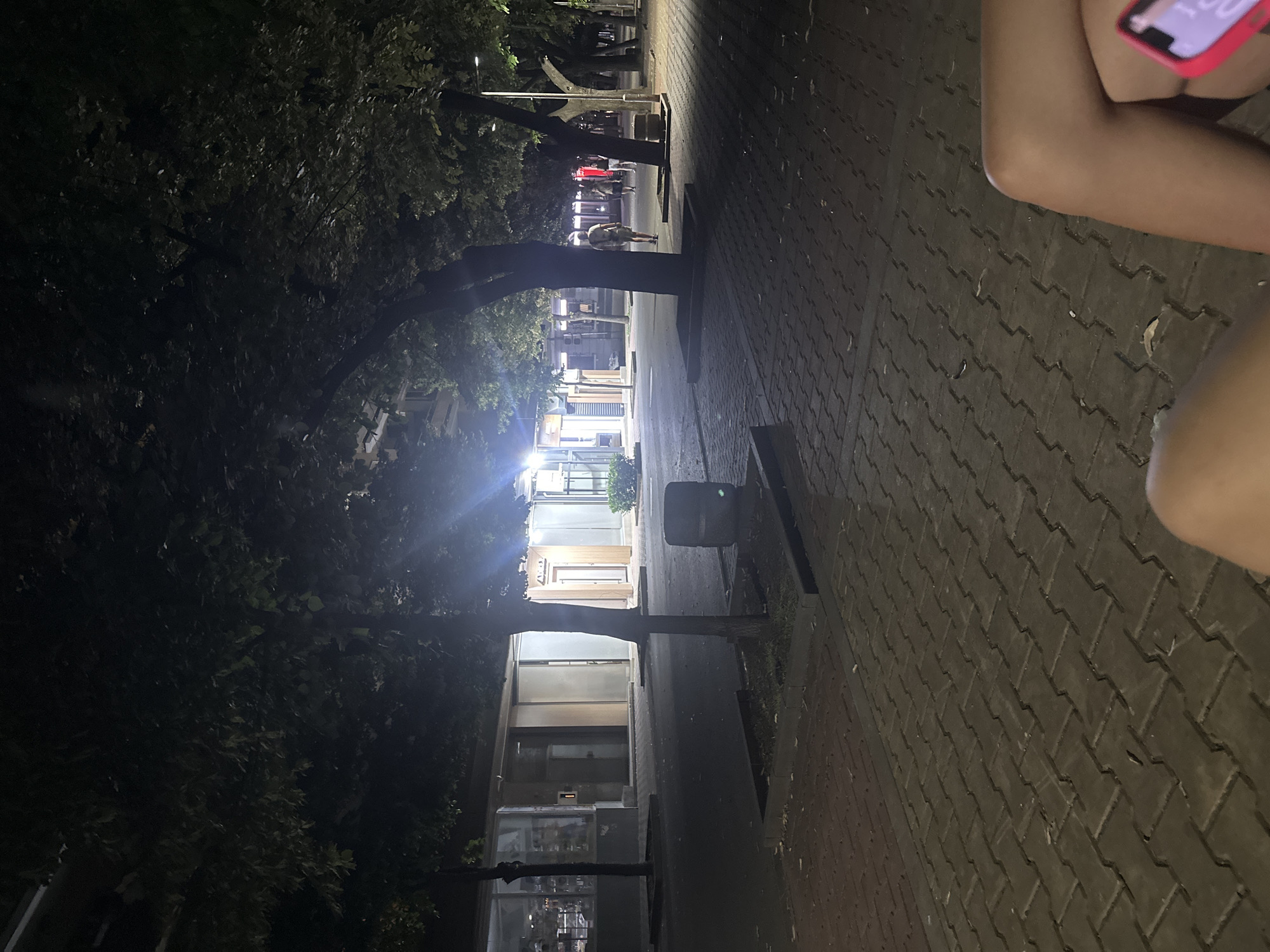 The image depicts a dimly lit outdoor scene, likely in the city of Shumen, Bulgaria, characterized by a paved walkway lined with trees. In the foreground, two legs are visible, suggesting the viewpoint of a person seated. The background shows several storefronts, some with illuminated interiors, and a few individuals walking along the pathway. Streetlights cast pools of light, illuminating sections of the pavement and the foliage of the trees. The overall atmosphere is tranquil and suggests an evening or nighttime setting.