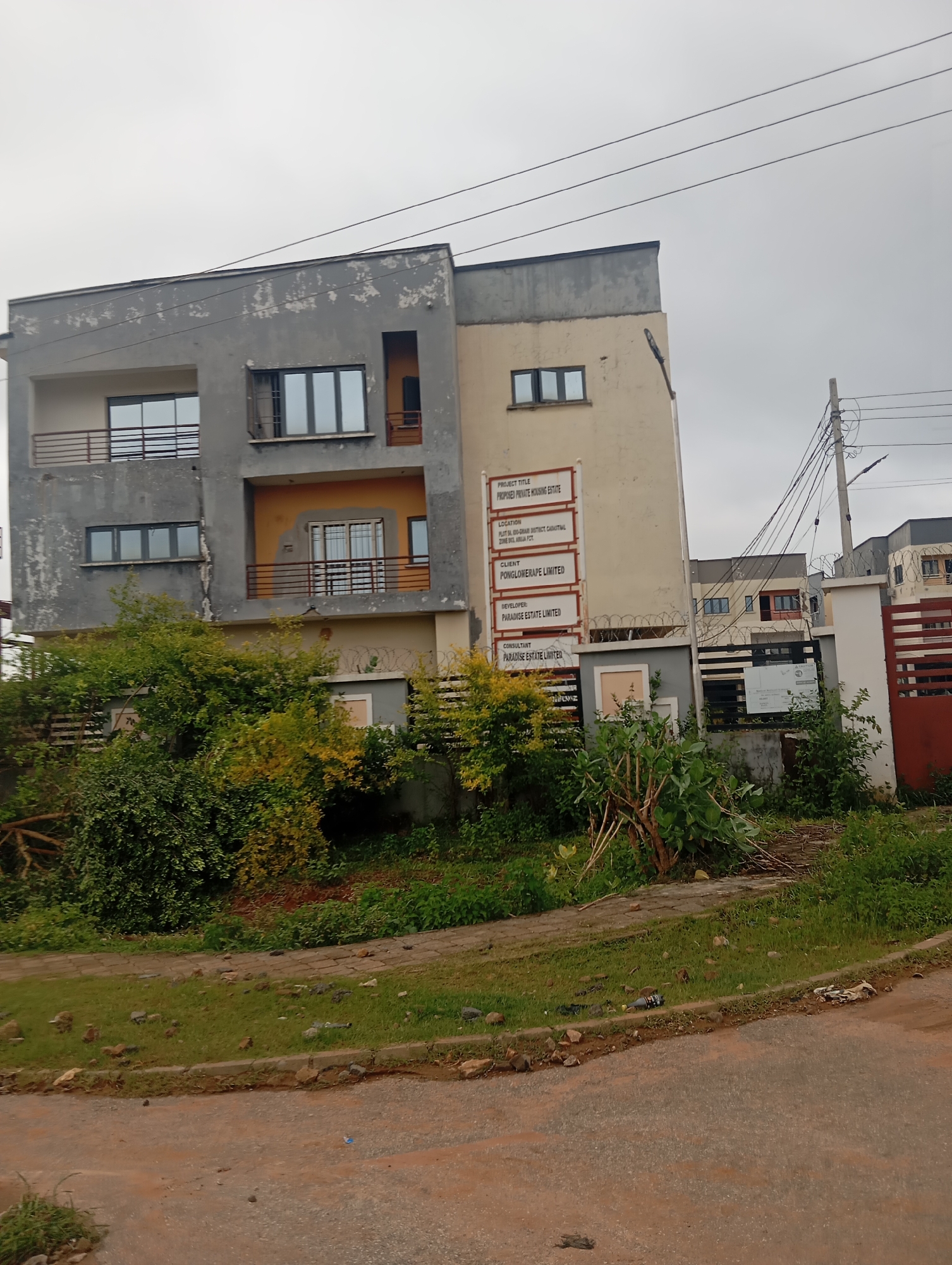 The image depicts a building under construction or renovation in Dutsen Alhaji, Nigeria. The building is a multi-story structure with a mix of grey and light yellow exterior paint, some of which appears to be peeling. The architecture features modern elements like large windows and balconies with railings. A prominent sign on the building lists project details, including "PROJECT TITLE: PROPOSED PRIVATE HOUSING ESTATE," "LOCATION: PLOT 781, B00-GHAR DISTRICT, DUTSEN-ALHAJI, GWARINPA, FCT," "CLIENT: PONGLOMERAPE LIMITED," "DEVELOPER: PARADISE ESTATE LIMITED," and "CONSULTANT: PARADISE ESTATE LIMITED." The surrounding environment includes lush green vegetation, a paved walkway, and a dirt road. Electrical wires and poles are visible, suggesting ongoing utility connections. The sky is overcast, indicating a cloudy day. No people are visible in the frame.