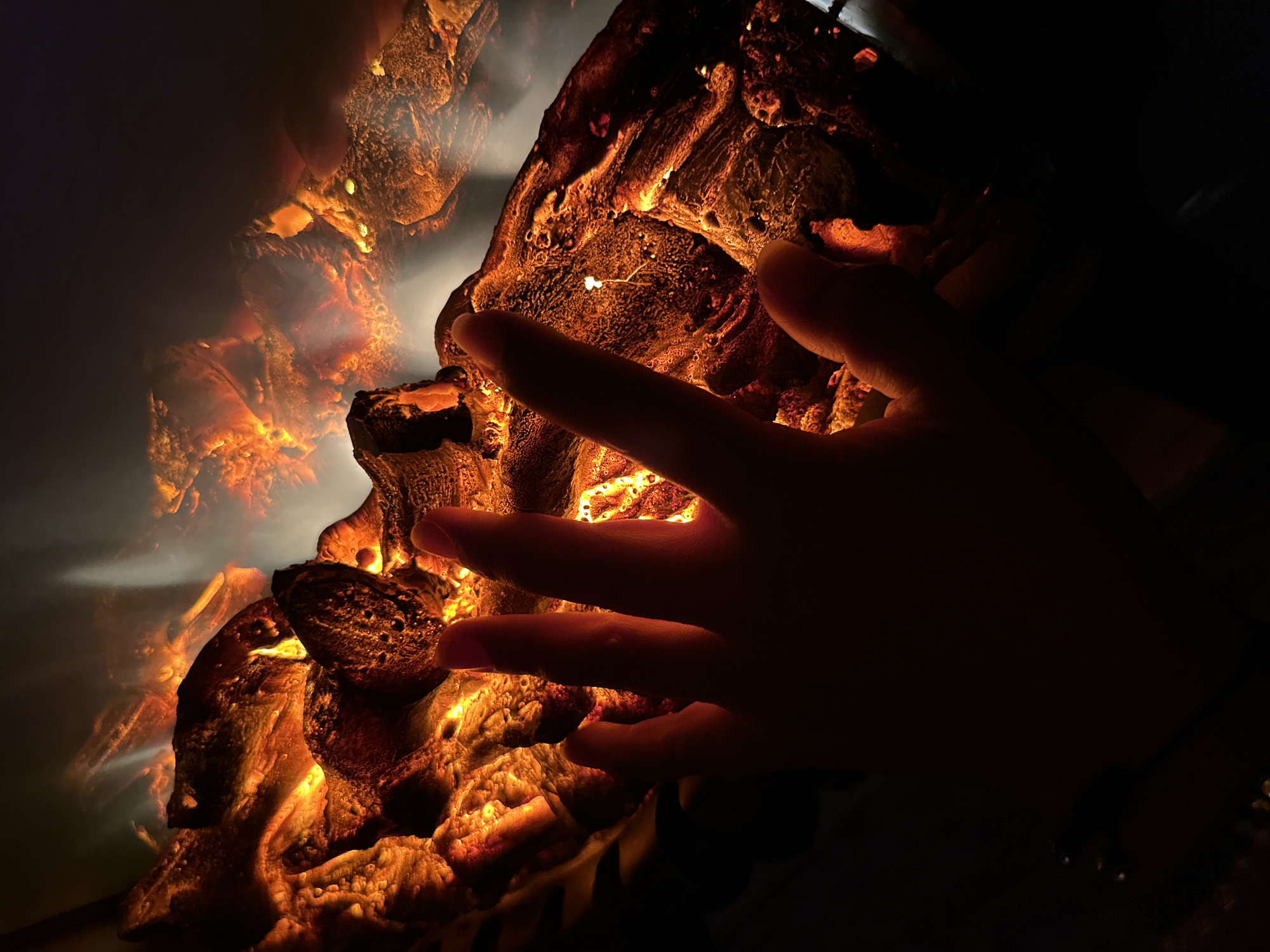A dimly lit image shows a person's hand in silhouette reaching towards a fiery, glowing artificial log set. The hand is positioned in the foreground, with its fingers extended and slightly curved. The artificial logs are a warm orange and yellow, with a textured surface that mimics embers and flames. Wisps of smoke or light appear to rise from the logs. The background is dark and out of focus, with some lighter areas that may indicate reflections or additional lighting. The overall impression is one of warmth and artificial fire. The location is unknown.