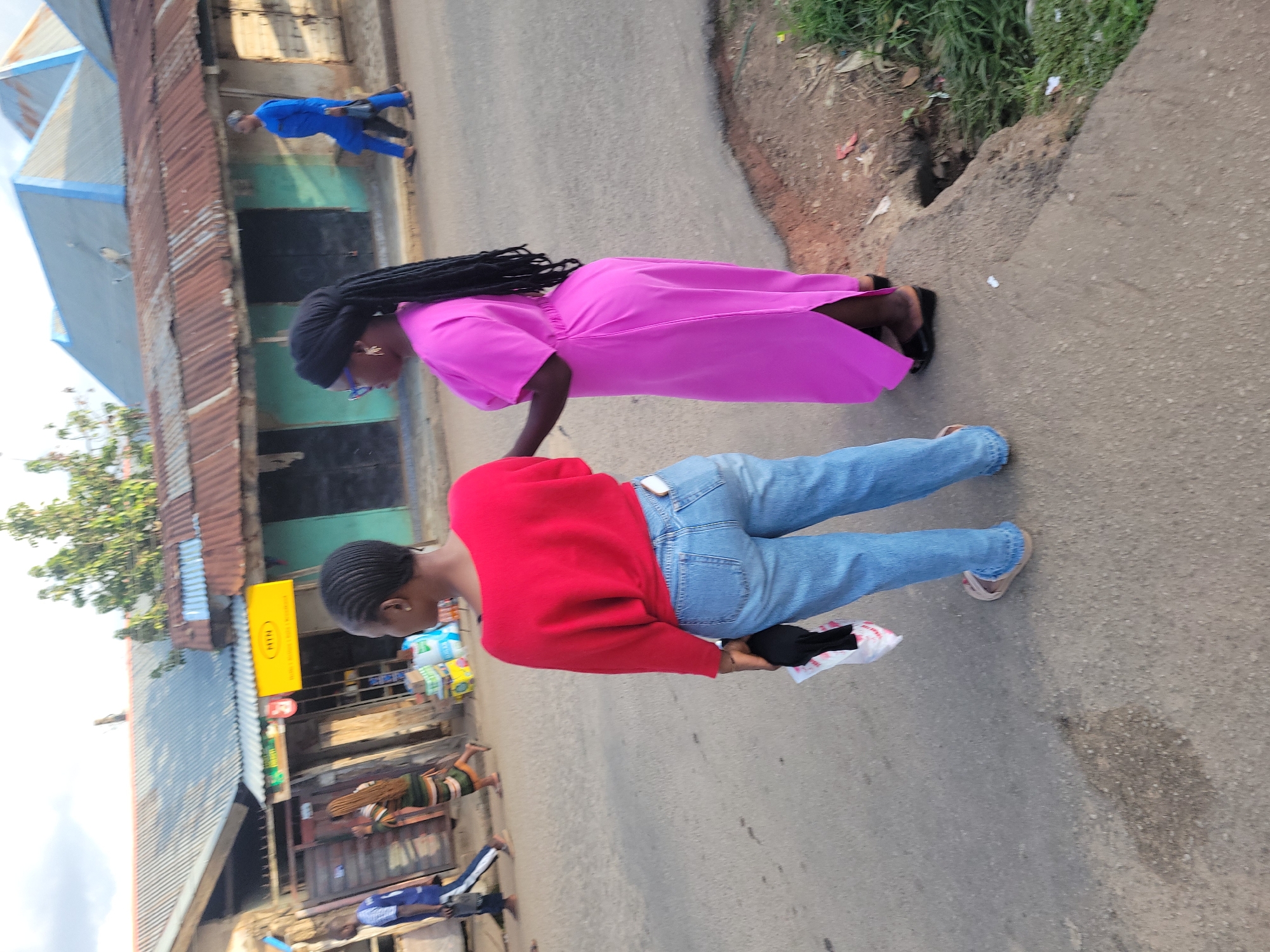 The scene depicts a street in Jos, Nigeria, during daylight hours, observed from a slightly elevated perspective behind two women in the foreground. The street surface is asphalt, with varying textures and patches.
In the immediate foreground, two individuals, identified as women, are oriented with their backs mostly towards the camera. The woman on the left wears a long-sleeved red top and light blue denim jeans with a light-colored sandal visible on her left foot. Her hair is styled in short, dark cornrows. She holds a white plastic bag containing a black item in her right hand. To her right stands another woman, dressed in a magenta-pink ankle-length dress with short sleeves and black wedge sandals. She has a dark headwrap, from which long, dark dreadlocks or braids cascade down her back. Her right arm is slightly bent at the elbow, and her hand is near the other woman's left arm. She appears to be wearing glasses with a purple frame and gold-colored hoop earrings.
In the midground and background, several other individuals are walking. On the far left, a man in a blue and white long-sleeved shirt and dark trousers walks away from the camera, carrying a dark bag. Further back and to the right of him, a woman with long braided hair is visible, wearing a vertically striped dress. On the far right, a man in a full blue traditional outfit, consistent with Nigerian attire, walks along the street.
The left side of the street is lined with commercial buildings, typically single-story structures with corrugated metal roofs of varying conditions; some are grey or blue, while others exhibit rust-brown hues. One building displays a prominent rectangular yellow sign with the text "MTN" at the top and additional smaller text below. Merchandise, including packaged goods and boxes, is visible outside some of these establishments. The street on the right edge transitions into a raised, unpaved verge with sparse green vegetation and exposed earth. A small, dark opening in the curb suggests a drainage point or culvert. The sky above is partially visible, appearing overcast or hazy.