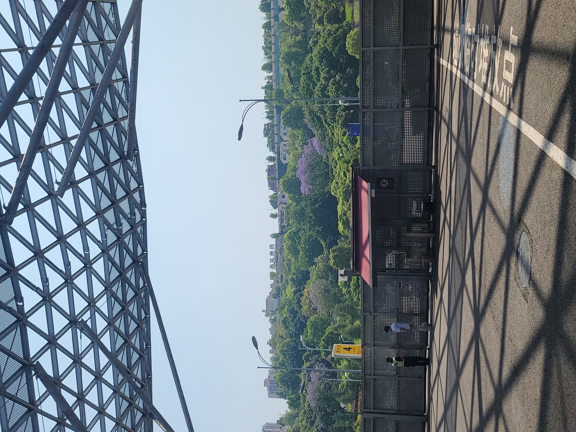 The image shows an outdoor scene in Liangjiang, China. The foreground features a complex metal structure, possibly part of a building or bridge. Adjacent to the structure is a bus stop with a shelter. Two people are present at the bus stop: one in a white shirt and light pants and another in a darker outfit, possibly security personnel. Beyond the bus stop, there is a dense area of trees with some visible buildings in the background. The road surface in the foreground has Chinese characters painted on it and is covered in the shadows cast by the metal structure. The sky is clear and blue.