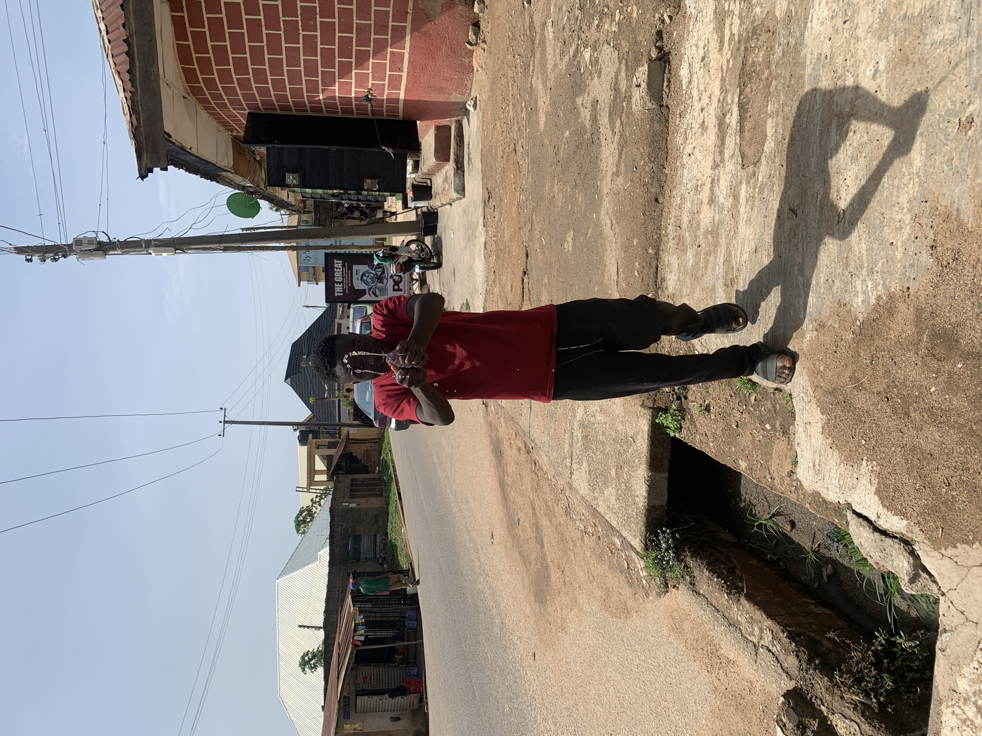 The image captures a full-body shot of a young man standing on a concrete roadside in what appears to be an urban or semi-urban setting in Jos, Nigeria, under bright daylight. The man, wearing a red short-sleeved t-shirt, dark trousers, and dark sandals, stands with his hands clasped together near his chest, looking towards the camera. He casts a long, distinct shadow on the dusty ground beside him, suggesting either early morning or late afternoon sunlight.
To his right (viewer's left), a paved road stretches into the distance, with what look like market stalls or small shops along its edge. To his immediate left (viewer's right), a concrete drainage ditch runs parallel to the road. This ditch contains some water, dirt, and scattered green vegetation.
In the background, a two-story building with a red brick facade and a corrugated metal roof is visible. Below this building, there's a power pole laden with numerous electrical wires and utility boxes, along with a green satellite dish attached to a lower part of the building. Several signboards are visible, one of which clearly displays the text "THE GREAT". A motorcycle is parked near the pole. Further in the background, other buildings with varying architectural styles, including a structure with a white corrugated roof, are visible under a clear, pale blue sky.
The scene depicts a typical street environment in the region, characterized by its infrastructure, building materials, and the strong tropical sunlight. The man is the central figure, actively posing for the photograph.