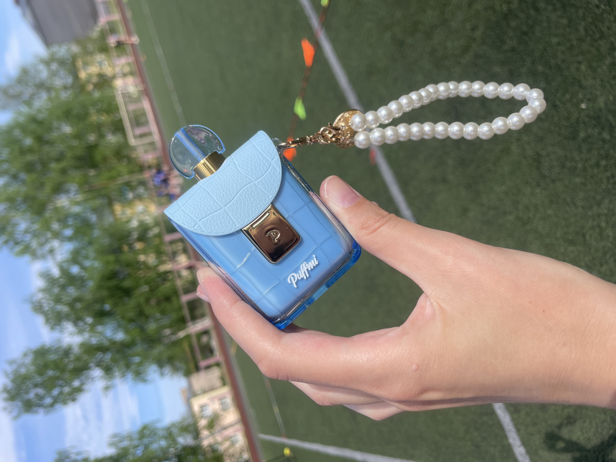 A close-up shot depicts a person's hand holding a small, light blue, purse-shaped bottle labeled "Puffmi." The bottle has a transparent top and a faux leather flap with a gold-colored clasp. A string of imitation pearls is attached to the purse. The background is blurred, showing a soccer field with white lines and colorful flags, along with trees and buildings. While the image was taken outside, there is nothing to indicate it was taken in Zav'yalovo, Russia.