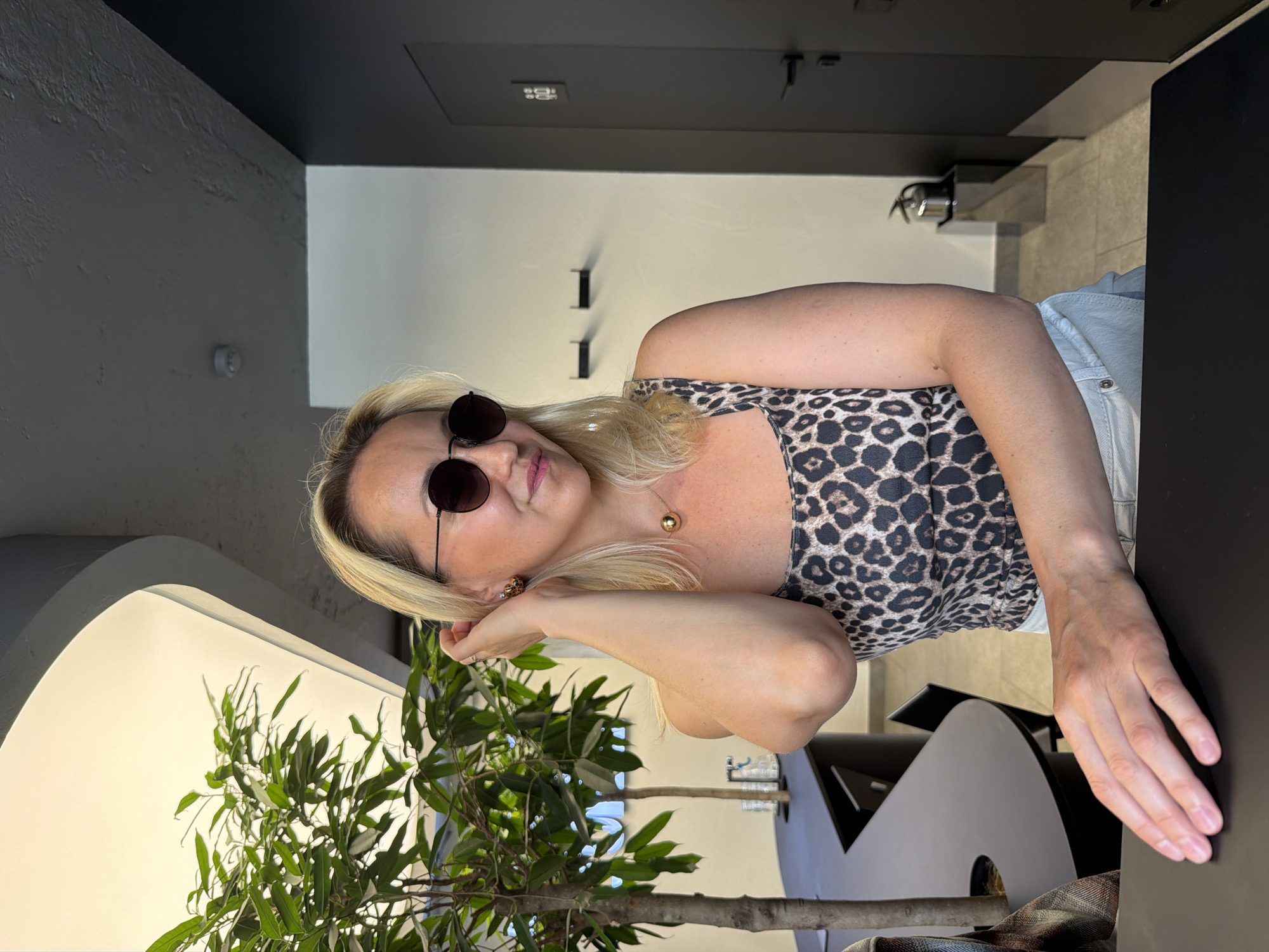 A woman with blonde hair, wearing round sunglasses, a leopard print top, and light-colored pants, is posing for a photograph indoors. She has her right hand raised to her hair and her left hand resting on a dark surface in front of her. The environment appears to be a modern establishment, possibly a cafe or office space, with a large plant on the left, dark furniture, and a door with a symbol on the right. There is no information in the image to suggest the location is in Izhevsk, Russia.