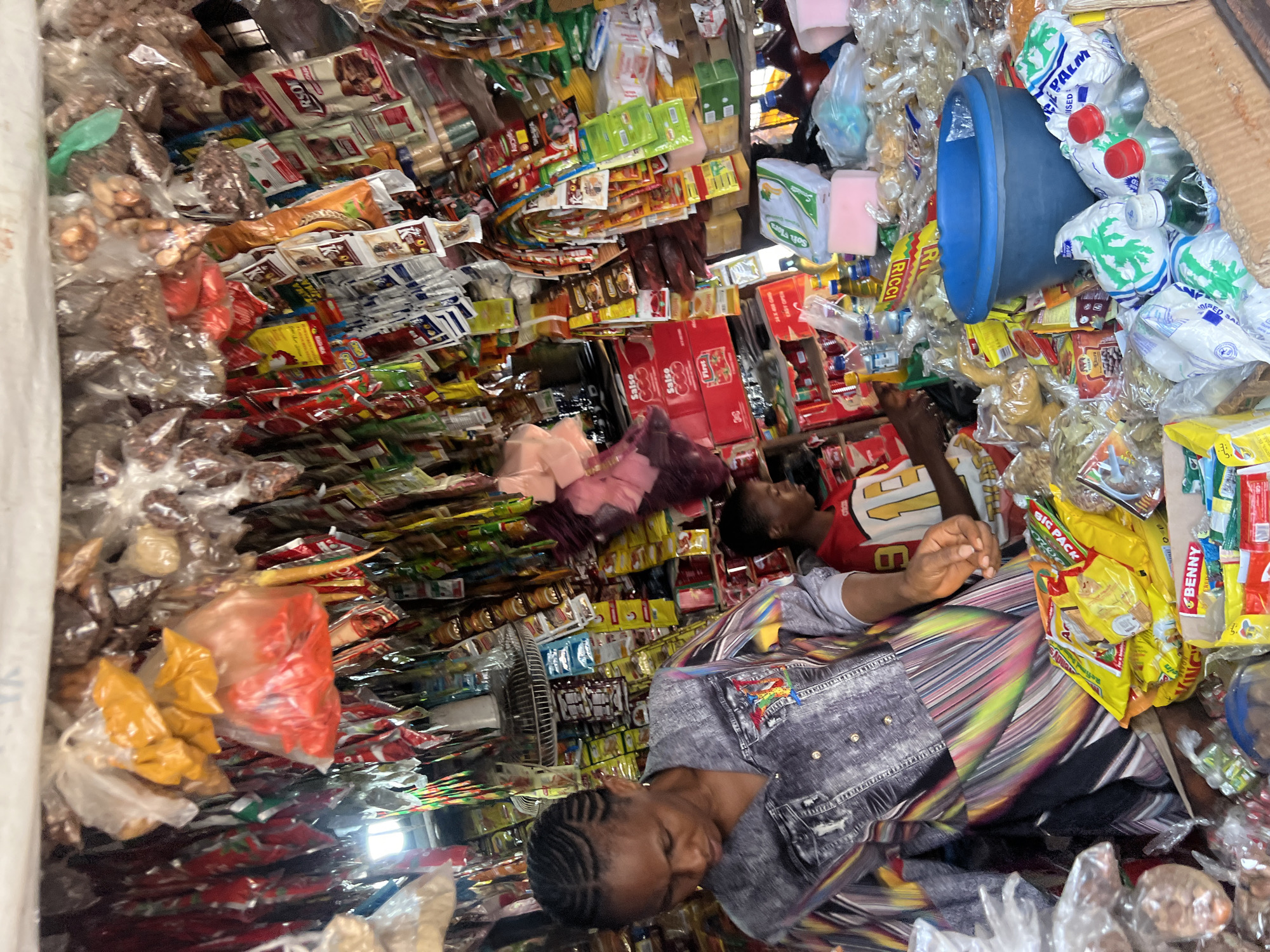 The image depicts a street market stall in Nkpor, Nigeria, overflowing with a wide variety of goods. The stall is densely packed with products ranging from food items, such as spices in plastic bags and packaged snacks, to bottles of liquids and what appear to be detergents. The items are hung from the ceiling and stacked on shelves, creating a visually busy environment. Two individuals are visible: a woman in the foreground, wearing a grey and colorful patterned shirt, appears to be attending to the stall, and a younger person is in the background wearing a red and white shirt. The lighting suggests it is daytime, with natural light filtering into the stall. The overall impression is one of a vibrant and active marketplace.
