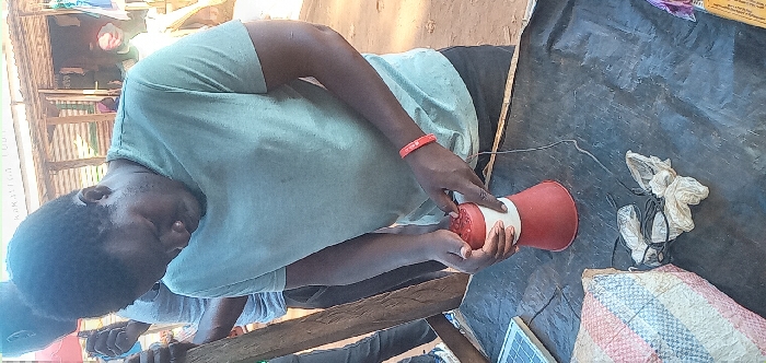A young man is standing in front of a wooden structure, wearing a light blue t-shirt and a red bracelet on his left wrist. He is looking down at a red and white object in his hands, likely a speaker. There is a black cable connected to the speaker and a solar panel on the table. The man is holding a speaker in front of a wooden structure, possibly a shop or a home. The scene takes place in Lwandeti, Kenya, a rural area characterized by simple structures and natural surroundings. The man is likely setting up the speaker for some purpose, possibly for music or announcements. The scene suggests an informal setting in a rural part of Lwandeti, Kenya.
