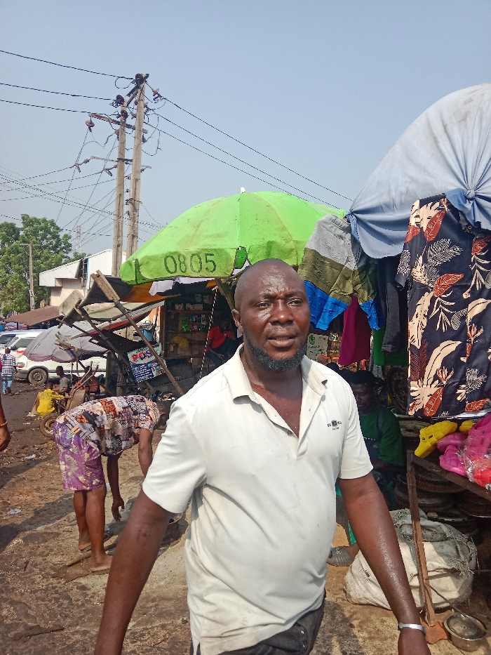 This image depicts a bustling outdoor market scene in Onitsha, Nigeria, under clear daylight.
In the immediate foreground, a dark-skinned adult male with a bald head and a short beard is positioned slightly off-center, facing towards the viewer with a neutral expression. He wears a light-beige short-sleeved polo shirt and dark shorts. Sweat is visible on his forehead, face, and arms. A simple silver-colored bracelet adorns his left wrist.
Behind this individual, the scene transitions into a dynamic market environment. To the left, another individual, wearing distinctive patterned shorts (purple, white, brown, and black) and barefoot, is bent over, engaged in an activity on the unpaved, uneven ground. Further left, a white van or bus is partially visible amidst other indistinct figures and market structures. A man in a yellow shirt is seated behind a small market cart.
To the right of the foreground figure, several market stalls are present. One prominent stall features a bright green umbrella with the number "0805" inscribed in white. Adjacent to it, another stall is covered by a light blue/grey umbrella. Clothing items, including a dark shirt with a light-colored leaf pattern, are displayed hanging from these structures. On a lower surface to the right, various items, notably bright yellow and pink molded plastic objects (possibly shoes or toys), are arranged for sale. A large, dirty white sack rests on the ground nearby. A partially obscured figure in a green shirt is visible behind the rightmost stall.
Overhead, multiple utility poles with a dense network of electrical wires crisscross the bright, clear sky. In the background on the far left, green foliage and trees are visible, along with parts of low-rise buildings. The ground throughout the market area appears to be unpaved, characterized by dirt and debris.