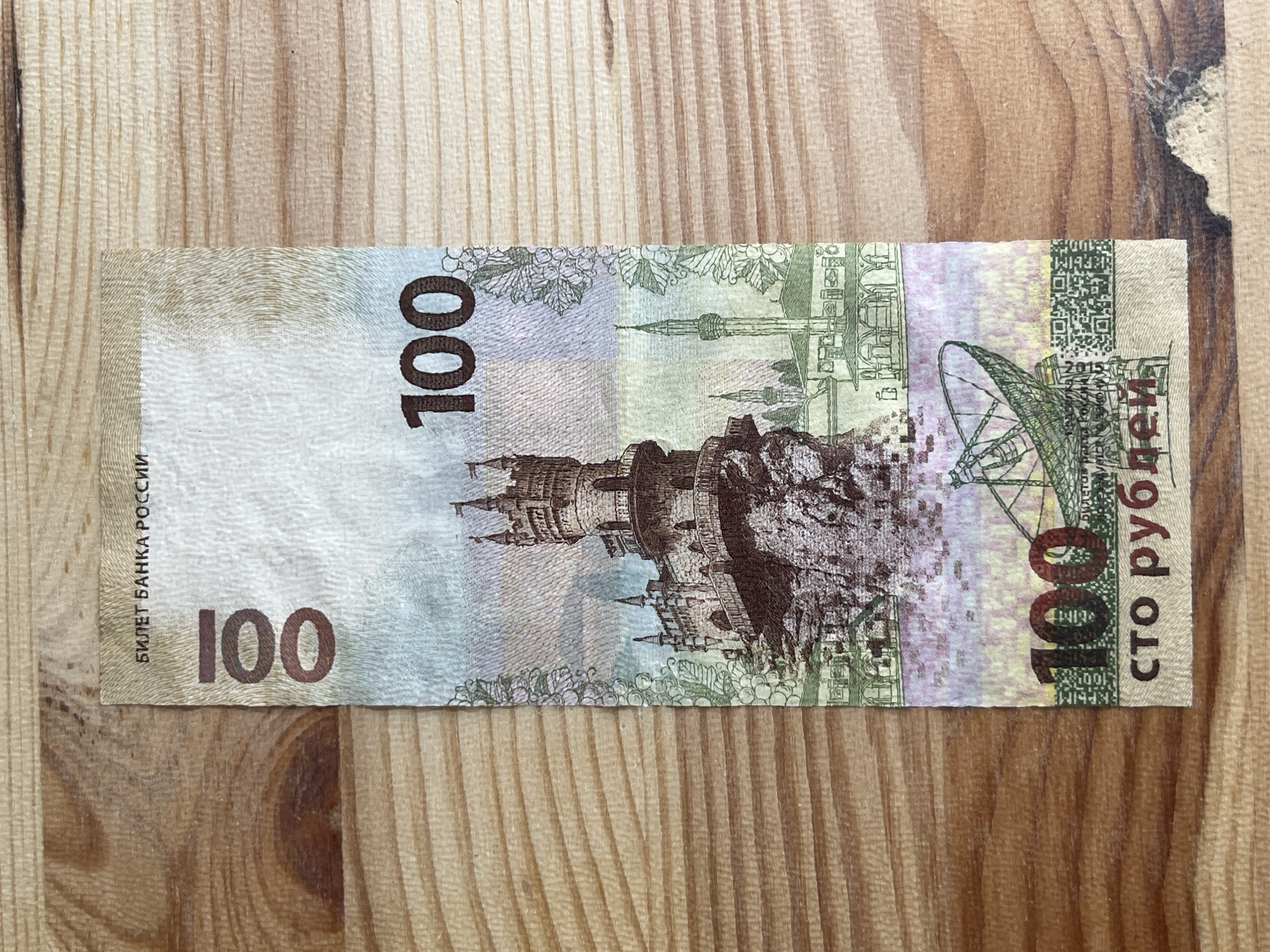 A 100-ruble banknote from Russia is shown on a wooden surface. The banknote features the Swallow's Nest castle in Crimea and the city of Sevastopol. The castle is perched on a cliff overlooking the sea, and the city skyline includes minarets and other architectural elements. The banknote is oriented vertically, with the denomination "100" printed in large numerals at the top and bottom. The text above the castle reads "БИЛЕТ БАНКА РОССИИ" (Bank of Russia Ticket). The text below the bottom "100" reads "СТО РУБЛЕЙ" (One Hundred Rubles). The banknote is slightly creased and shows some wear. The wooden surface appears to be a table or countertop, with visible grain and texture.