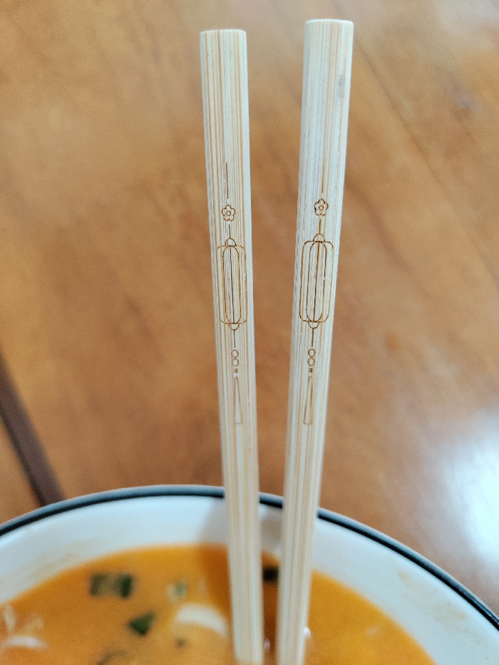The image displays a close-up, high-angle view of a meal in Thành phố Đà Lạt, Vietnam. A pair of light-colored bamboo chopsticks are positioned vertically within a white bowl accented with a black rim. Each chopstick is engraved with an identical design, featuring a small flower at the top, a central rectangular lantern motif, and a tassel-like element towards the bottom. The bowl contains a vibrant orange-red liquid, indicative of a soup or broth, with indistinct green and lighter-colored ingredients partially submerged. The background is an out-of-focus, warm-toned light brown surface, consistent with a wooden table. No individuals are visible in the scene.