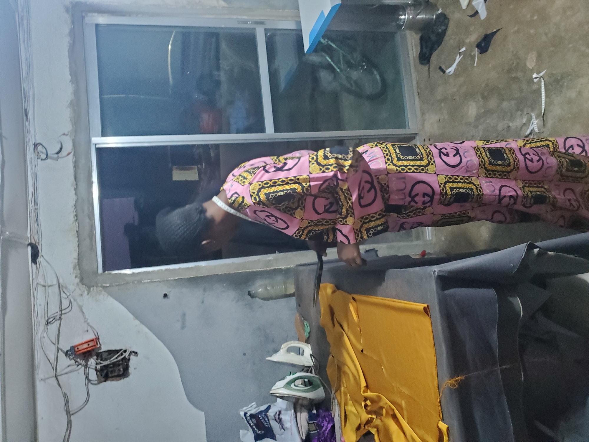 A person wearing a pink and gold patterned kaftan is leaning over a table, seemingly engaged in some activity. The person has dark skin and is wearing a dark cap. A measuring tape is wrapped around their neck, suggesting they might be a tailor or seamstress. The setting appears to be a workshop or a domestic space with construction elements. In the background, a window shows a reflection of a bicycle.
On the table, there are two irons, one white and one green and white, placed on top of a yellow fabric. A clear plastic bottle is also visible on the table. Various wires and an electrical junction box are mounted on the wall to the left, indicating ongoing or recent electrical work. The floor is rough concrete. The overall scene suggests a somewhat cluttered and utilitarian environment, possibly a tailoring shop in Jos, Nigeria. The lighting suggests it is daytime, but the interior lighting is the primary source. No weather cues are apparent. There is no visible text in the image.