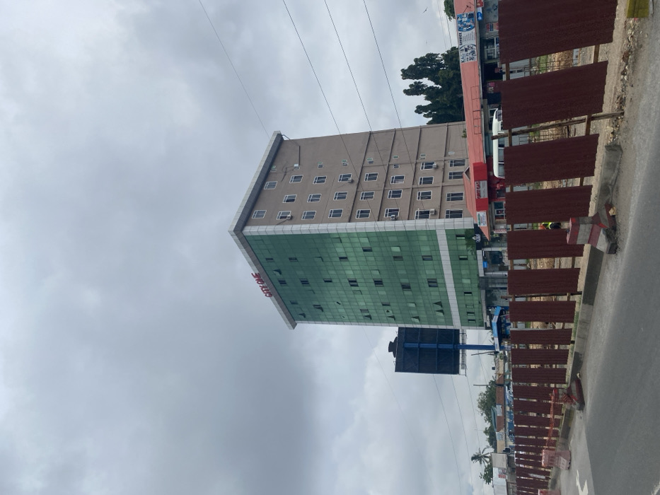 The image shows a tall building with a green glass facade and a sign that reads "CITY ONE". The building is located on the corner of a street, with a fence and several vehicles parked in front of it. There are several other buildings in the background, including a billboard with a blue background. The sky is overcast with clouds, and there are power lines running overhead. The image was taken in Dar es Salaam, Tanzania, at the coordinates -6.773, 39.241. The scene appears to be on a busy street in a commercial area, with a mix of modern and older buildings. There are no people visible in the image.