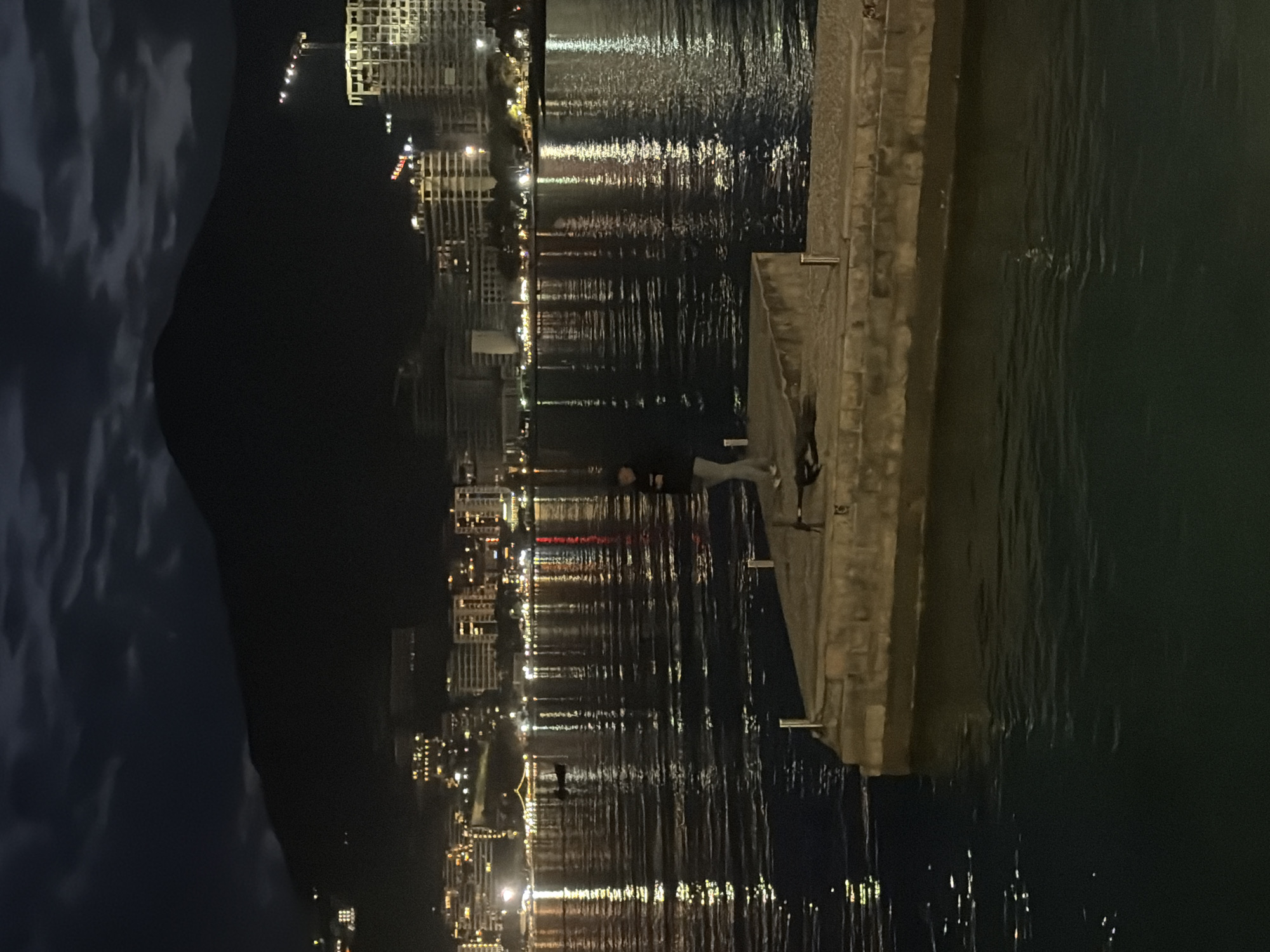 This nighttime image captures a tranquil coastal scene, likely in Budva, Montenegro, looking across a body of water towards an illuminated cityscape. The sky, dark but with visible cloud formations on the left, extends above the distant buildings.
In the foreground, a sturdy stone or concrete quay occupies the right side of the frame, extending into the water. On this quay, at least two figures are present. One individual, dressed in light-colored clothing (appearing as a white top and light trousers), is seated or leaning near the water's edge, their back mostly to the viewer, seemingly observing the view. Further back, another person in darker attire stands near a large object covered with a dark tarp, possibly a boat or equipment stored on the quay.
The central part of the image is dominated by the calm body of water, which vividly reflects the numerous artificial lights from the distant city. These reflections create elongated, shimmering streaks of yellow, white, and red across the water's surface, indicating a serene environment. The distant cityscape features various buildings, their lights forming a sparkling panorama against the dark night.
The scene conveys a peaceful atmosphere, with the individuals on the quay appearing to be quietly taking in the beautiful nocturnal view of the illuminated city reflecting on the water. No visible text is present in the media file.