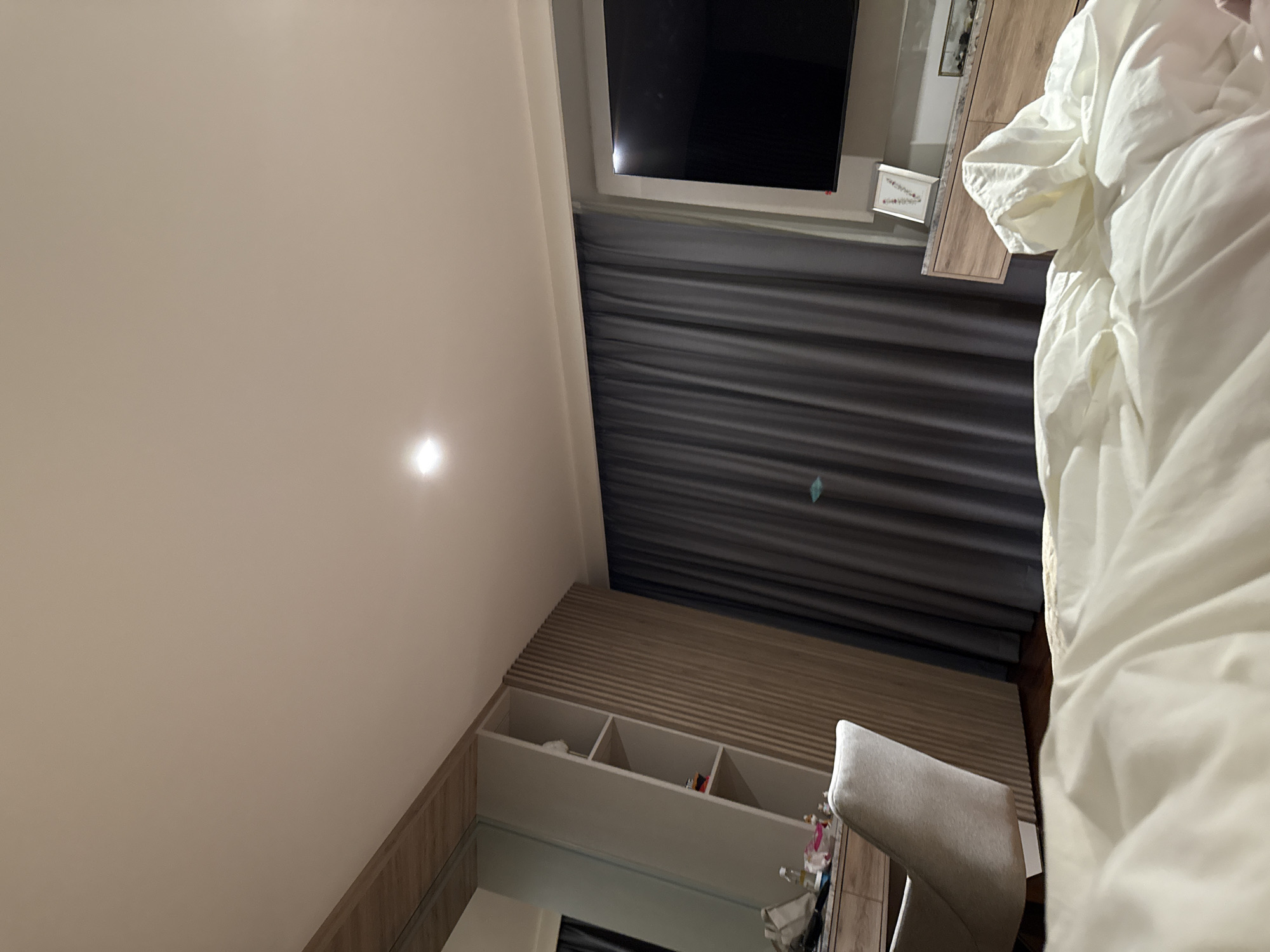 The image depicts a bedroom interior. In the foreground, a rumpled white duvet is visible, suggesting a bed. To the left, a light-colored upholstered chair is positioned in front of a wooden dresser with drawers. Behind the chair, a wall unit with shelves is present, and a large mirror covers a portion of the wall. Further back, dark gray curtains hang across a window. To the right, a large flat-screen television is mounted on the wall above a wooden cabinet. A framed picture with floral motifs is placed on the cabinet. The room is lit by a ceiling light fixture. The location is identified as San Bernardino Tlaxcalancingo, Mexico.