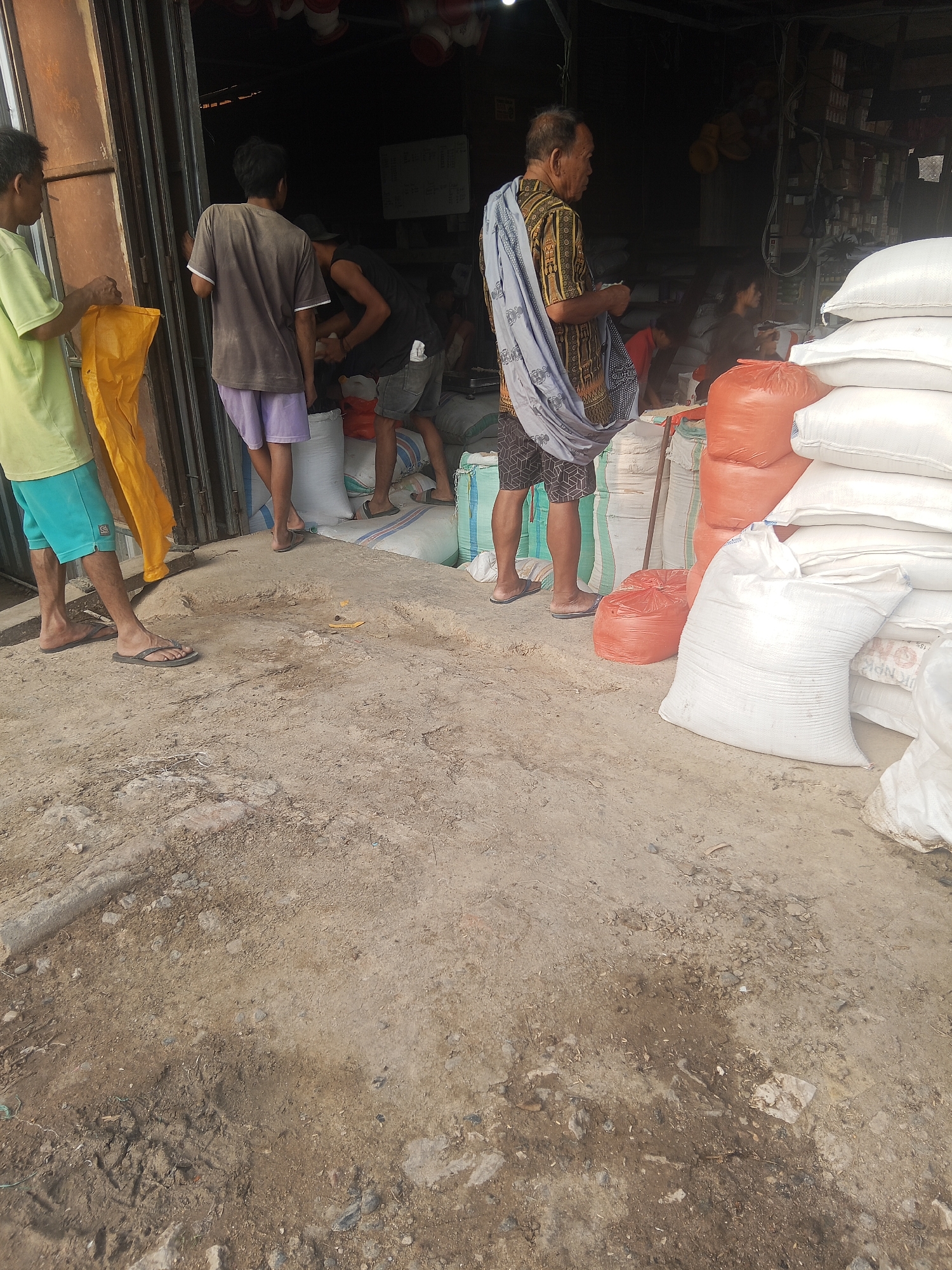 The image depicts an outdoor scene in what appears to be a marketplace or storage area, likely in Batu Papan, Indonesia. The ground is made of dirt and debris, with some areas showing signs of dampness or mud. Several individuals are present, engaged in various activities. On the left, a person wearing a light green t-shirt and blue shorts is holding a yellow bag. Next to them, another person in a dark grey t-shirt and purple shorts stands near a pile of white sacks. Further in the background, another individual in a black tank top and grey shorts is interacting with someone behind a counter. An older man, wearing a patterned shirt with a light blue draped cloth over his shoulder and patterned shorts, is standing near large sacks, some of which are orange and white. The background shows the interior of what could be a shop or warehouse with shelves and merchandise. The overall impression is of a working environment where goods are being handled or stored.