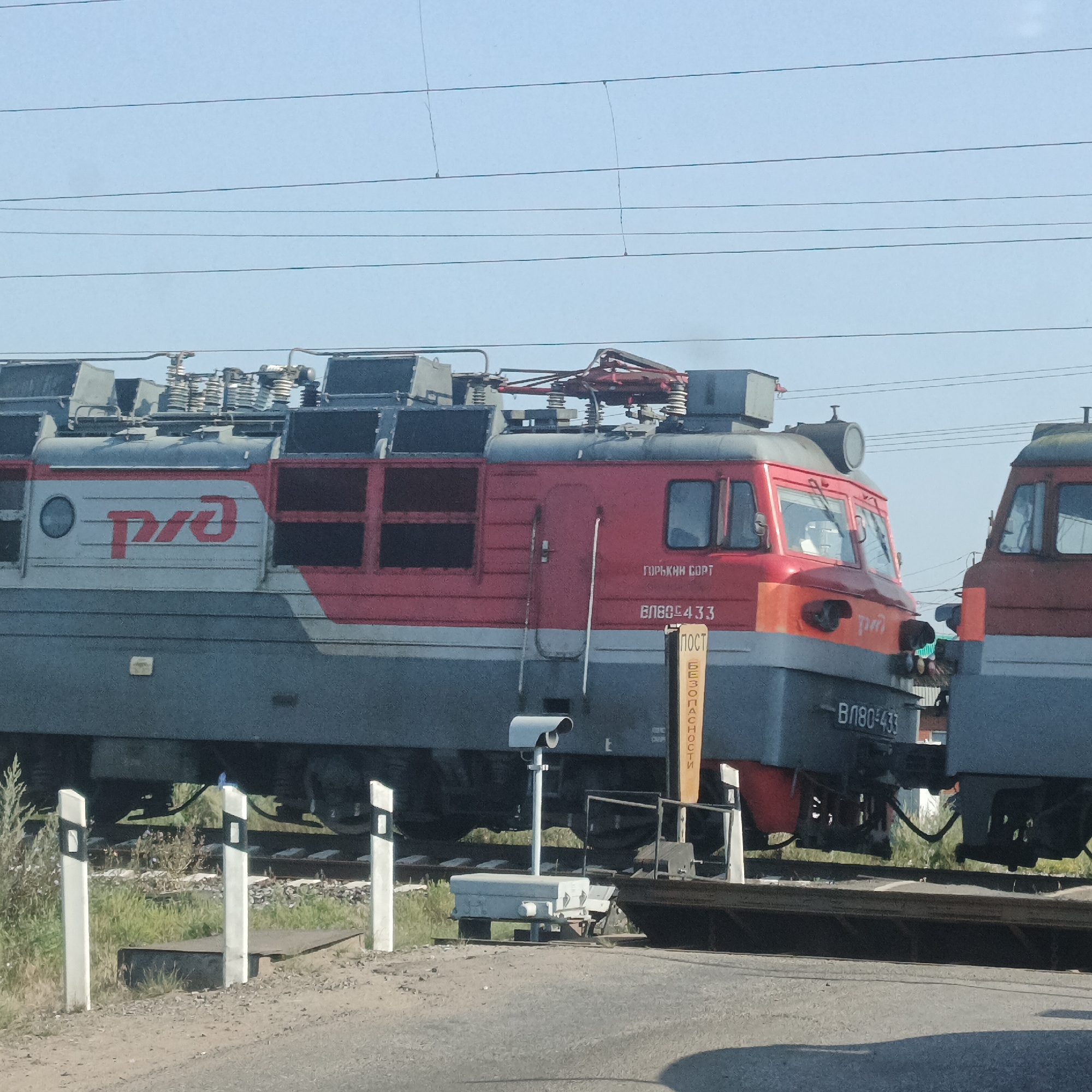 An exterior medium shot captures two red and gray electric locomotives of Russian Railways on a bright, clear day. The locomotive on the left is closer to the viewer and features the Russian Railways logo, a red circular emblem with stylized "RZD" in white. The number "ВЛ80-433" is visible on its side. The locomotive on the right is partially visible, situated to the right and slightly behind the first. Both locomotives are positioned on railway tracks, with overhead power lines visible against the sky.
In the foreground, a road crosses the railway tracks. Several white and black striped road markers, typical of level crossings, are positioned along the road. A security camera is mounted on a pole near the tracks. A yellow sign with vertical black lettering, reading "ПОСТ БЕЗОПАСНОСТИ" (POST BEZOPASNOSTI, meaning "Safety Post"), is also present. The ground is a mix of asphalt and gravel, with patches of green grass and vegetation on either side of the tracks. The scene is likely at a railway crossing in the city of Kambarka, Russia, given the context. No people are visible in the image.
