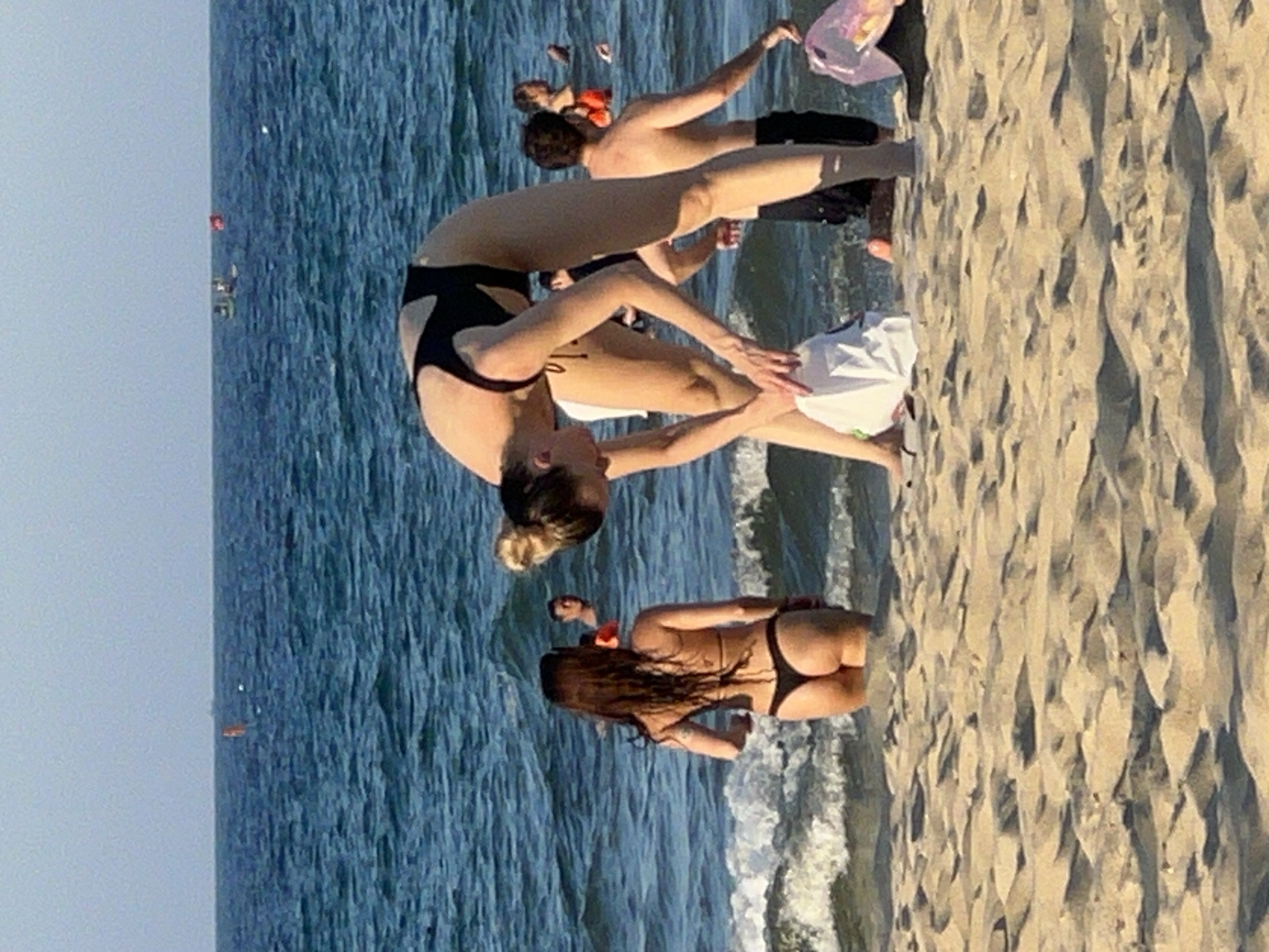 The image depicts a beach scene in Da Nang, Vietnam, with several individuals in the water and on the sand. In the foreground, on the sandy beach, a person wearing a black swimsuit is bent over, holding a white bag. To their left, another person in a black bikini stands in the shallow water, facing away from the camera. In the background, several other people are visible in the ocean, some with buoys. The water is blue with moderate waves, and the sky is a pale blue. The scene appears to be a casual day at the beach.