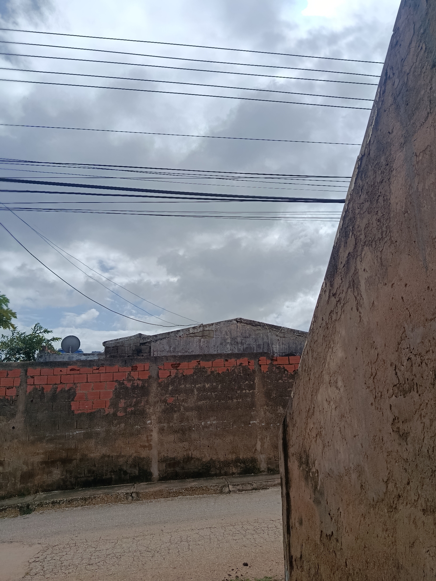 An outdoor, eye-level, medium shot captures a cloudy sky with visible overhead power lines. In the foreground, a textured, light brown wall extends from the right edge of the frame towards the center. To the left, a lower, weathered brick wall is visible, partially covered in darker grime and patches of missing mortar, revealing the red brick underneath. Beyond the brick wall, the top of a building with a sloped, dark roof is seen, and a small satellite dish is mounted on the roof. A tree with green foliage is to the far left. A cracked asphalt road runs horizontally across the bottom of the frame. The scene appears to be a residential or urban environment in Juan Griego, Venezuela. There are no people or animals visible.