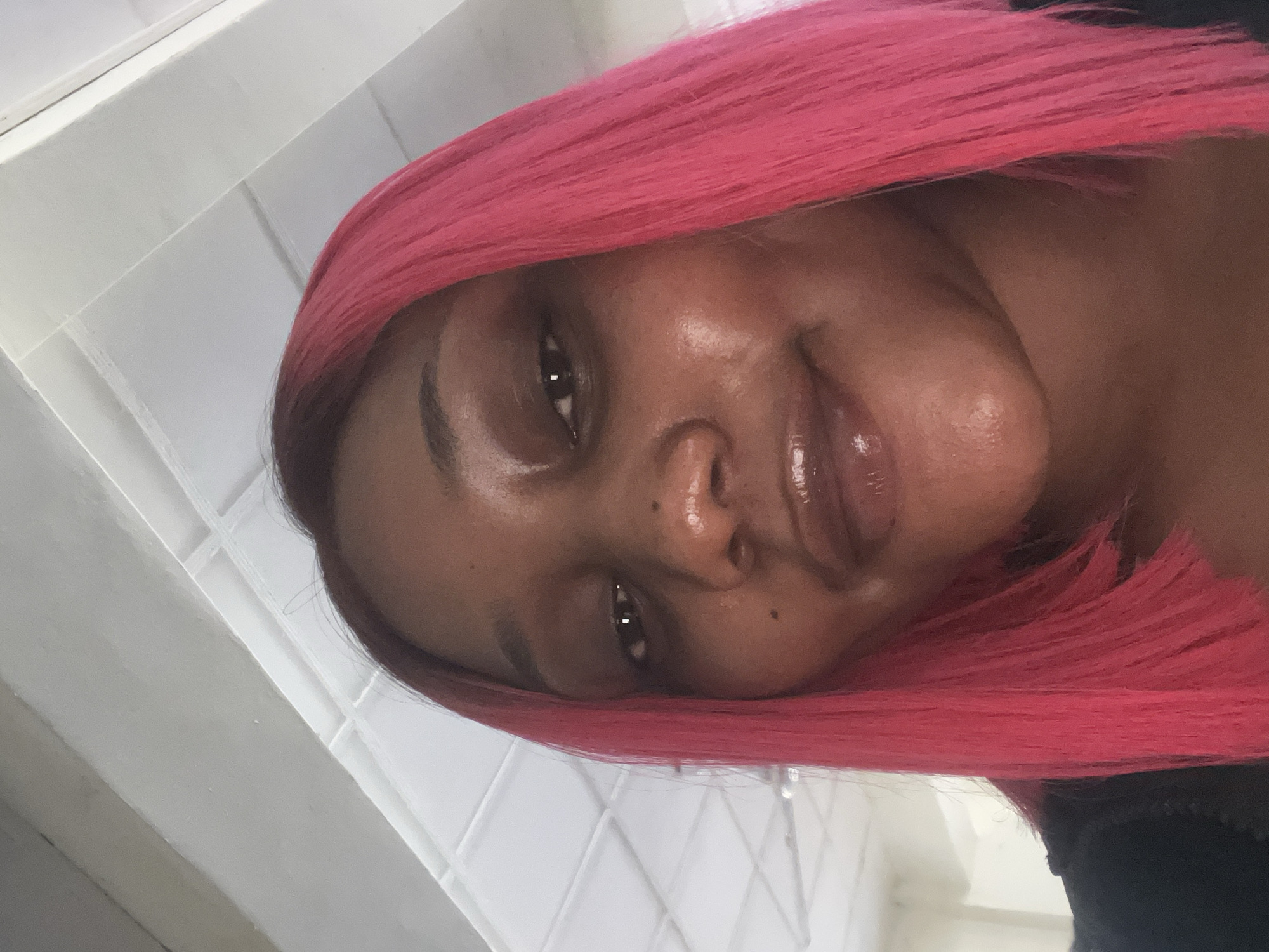 A close-up selfie of a young Black woman with bright pink hair. She is looking directly at the camera with a neutral expression. Her face is slightly angled, and the lighting suggests an indoor environment, possibly a residential or office space, given the textured white ceiling in the background. There are no specific landmarks visible that would indicate a location in Jos, Nigeria.