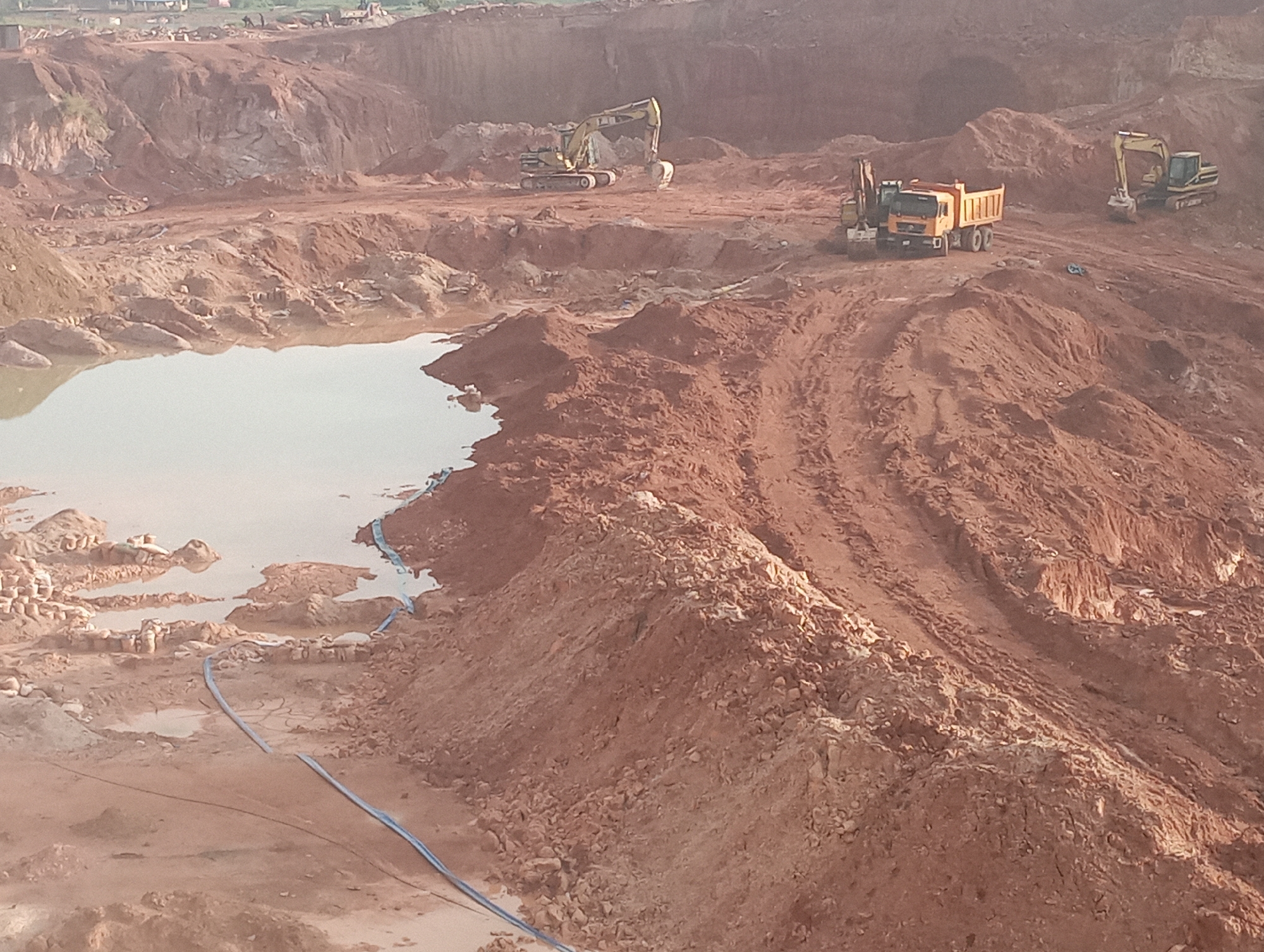 The image depicts an active construction site, likely a quarry or large excavation project, in Kwang, Nigeria. The terrain is characterized by large mounds of reddish-brown earth and exposed rock faces, indicating significant earthmoving operations. In the foreground, a body of murky water has accumulated, reflecting the overcast sky. Several pieces of heavy machinery are visible, including excavators and a large dump truck, suggesting ongoing excavation and material transport. A blue hose runs along the ground, possibly for water management. The background shows more layered earth and distant structures, hinting at the scale of the development. The scene is devoid of people, emphasizing the mechanical nature of the work.