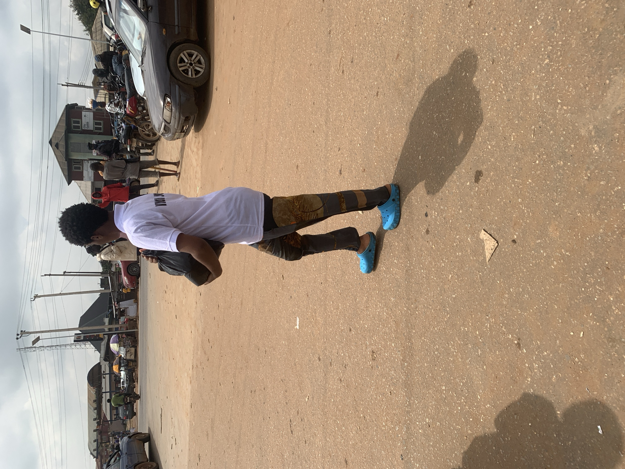 The image captures a street scene in Shagamu, Nigeria, on a cloudy day. A person wearing a white polo shirt with black lettering that reads "VIVO SONG" is walking from left to right across a dirt road. They are carrying a black plastic bag in their right hand and wearing blue Croc-style shoes. Their pants are dark gray with a pattern of tan and brown abstract designs. To the right of the person, a dark gray sedan is parked on the street. In the background, several motorcycles are parked, and there are buildings with corrugated metal roofs and a few people standing around. Further in the distance, more vehicles and market stalls are visible, suggesting a busy commercial area. Power lines crisscross the sky overhead. The ground is dusty and uneven, with scattered debris.