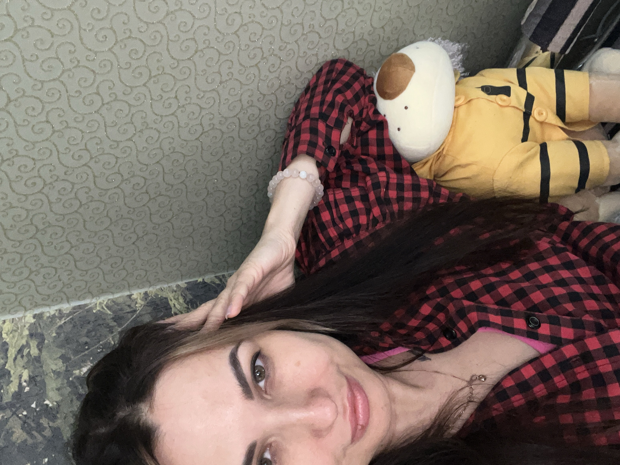 An overhead shot captures a woman lying down, looking directly at the camera with a subtle, relaxed smile. Her long, dark brown hair frames her face, and her right hand is casually placed near her head. She wears a red and black plaid button-up shirt, partially open to reveal a pink garment underneath. A light-colored, possibly rose quartz, beaded bracelet adorns her right wrist, and a delicate silver necklace with a small pendant is visible around her neck. Her makeup includes defined eyebrows, eyeliner, and light pink lip color.
Next to the woman, partially obscured, lies a white plush toy with a distinct brown spot on its head and small dark features. The toy is dressed in a yellow shirt featuring black horizontal stripes and black buttons, resembling a bumblebee or similar character.
The background consists of two distinct surfaces. Behind the woman and the toy is a wall covered in light-colored wallpaper with a shimmering, swirling, abstract pattern. Below her head and to the left, a darker surface with a marbled, organic pattern is visible, possibly a floor or another piece of furniture. The lighting is soft and even, suggesting an indoor setting. There is no visible text in the image. The scene conveys a calm and casual atmosphere.