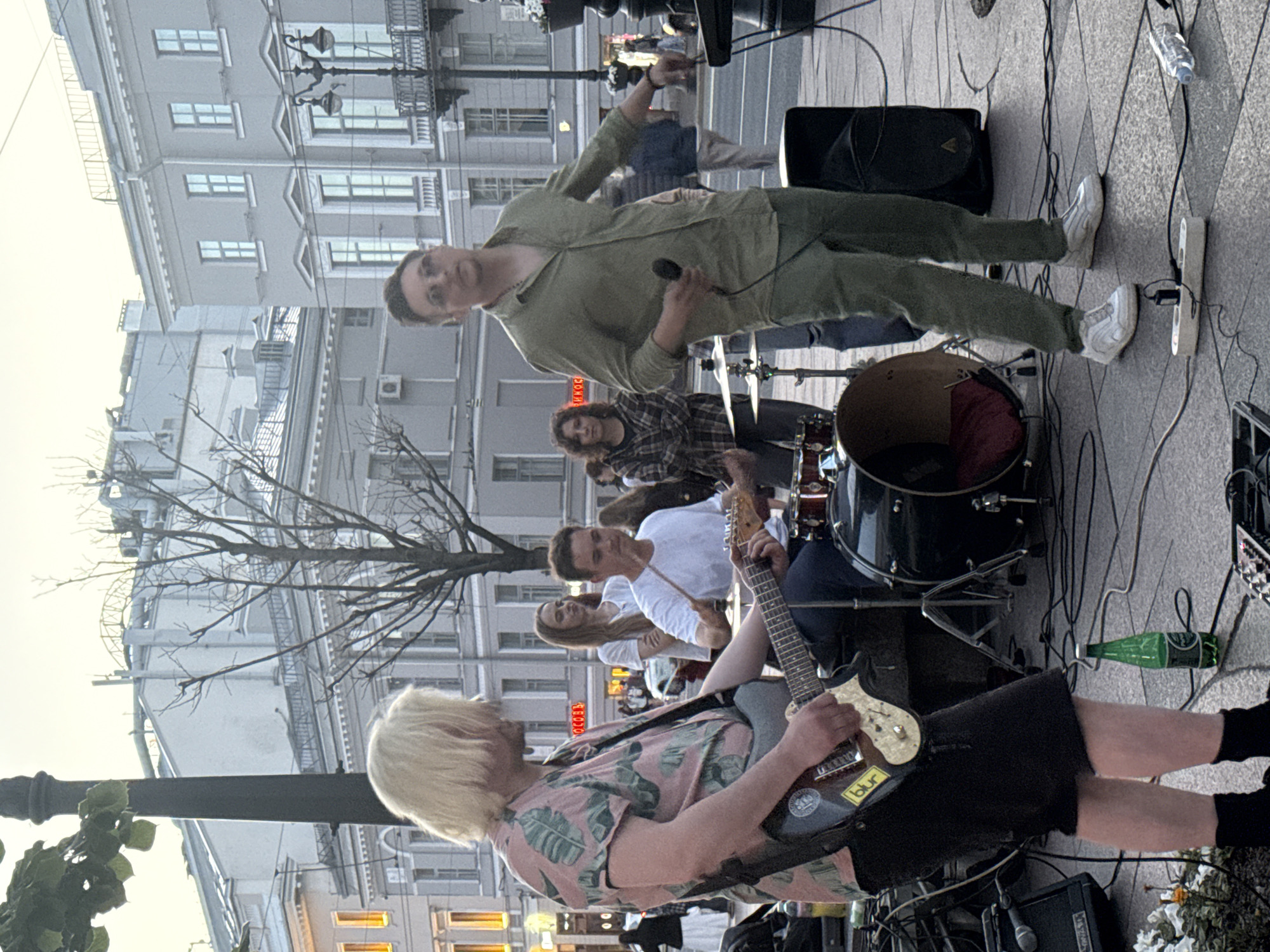 A band is performing live on a street in Saint Petersburg, Russia. The band members are playing guitars and drums. The lead singer is wearing a green shirt and pants and holding a microphone. A guitarist in the foreground has blond hair and is wearing a pink shirt with a leaf pattern. The scene appears to be set up on a paved area, possibly a sidewalk or square, with buildings in the background. The lighting suggests it is late afternoon or early evening, with a soft, diffused light. The mood seems to be casual and energetic, typical of a street performance. A green bottle of what appears to be beer is on the ground near some electrical equipment. There is some visible text on a sticker on the guitar that says "blur".