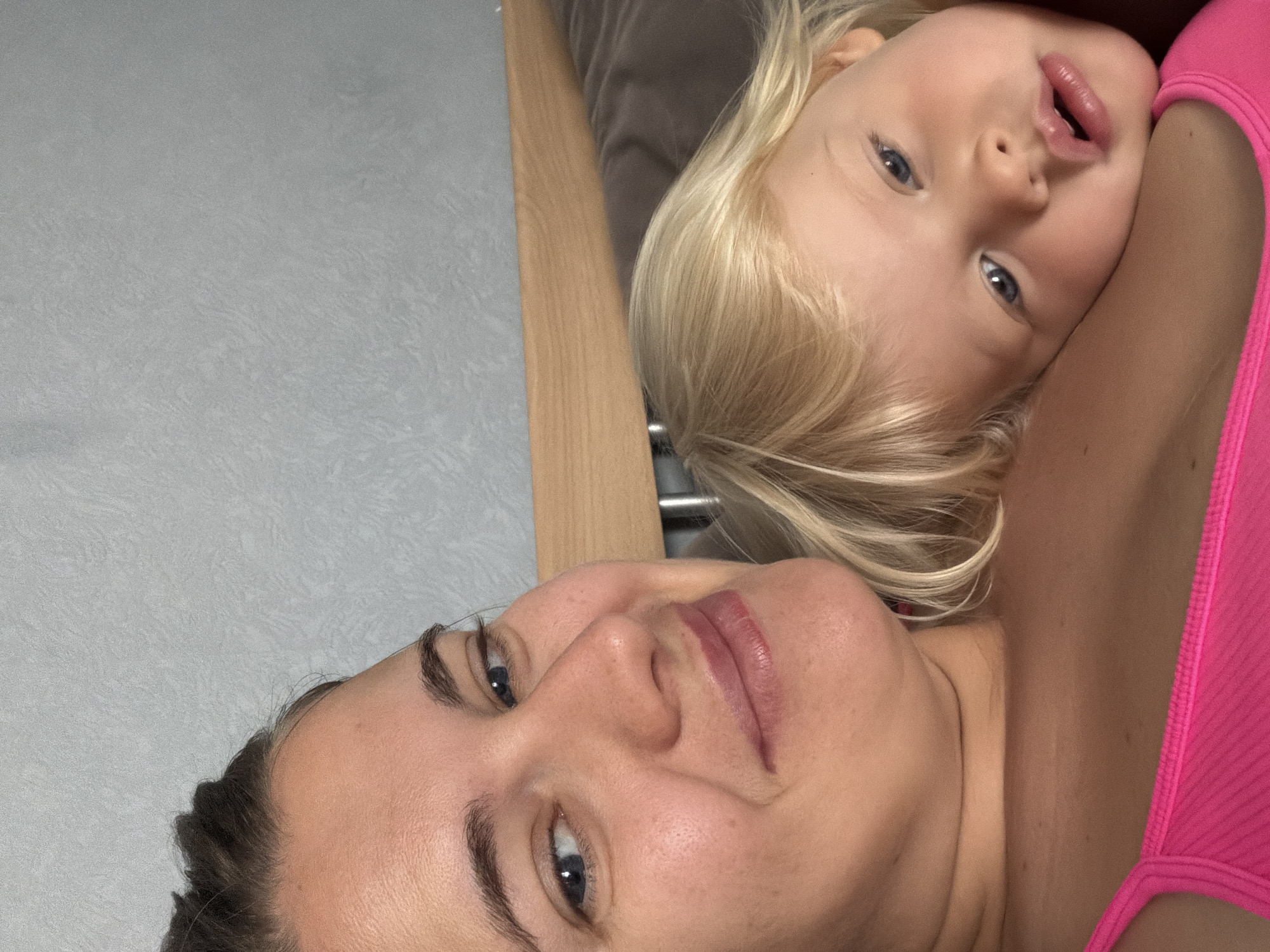 The image shows a close-up of a woman and a young child. The woman has dark hair pulled back and is looking towards the camera with a slight smile. The child has blonde hair and is resting their head on the woman's shoulder, looking away from the camera. They are both indoors, with a textured gray wall and a wooden headboard visible in the background. The scene does not contain any elements that would indicate the location is in Sochi, Russia.