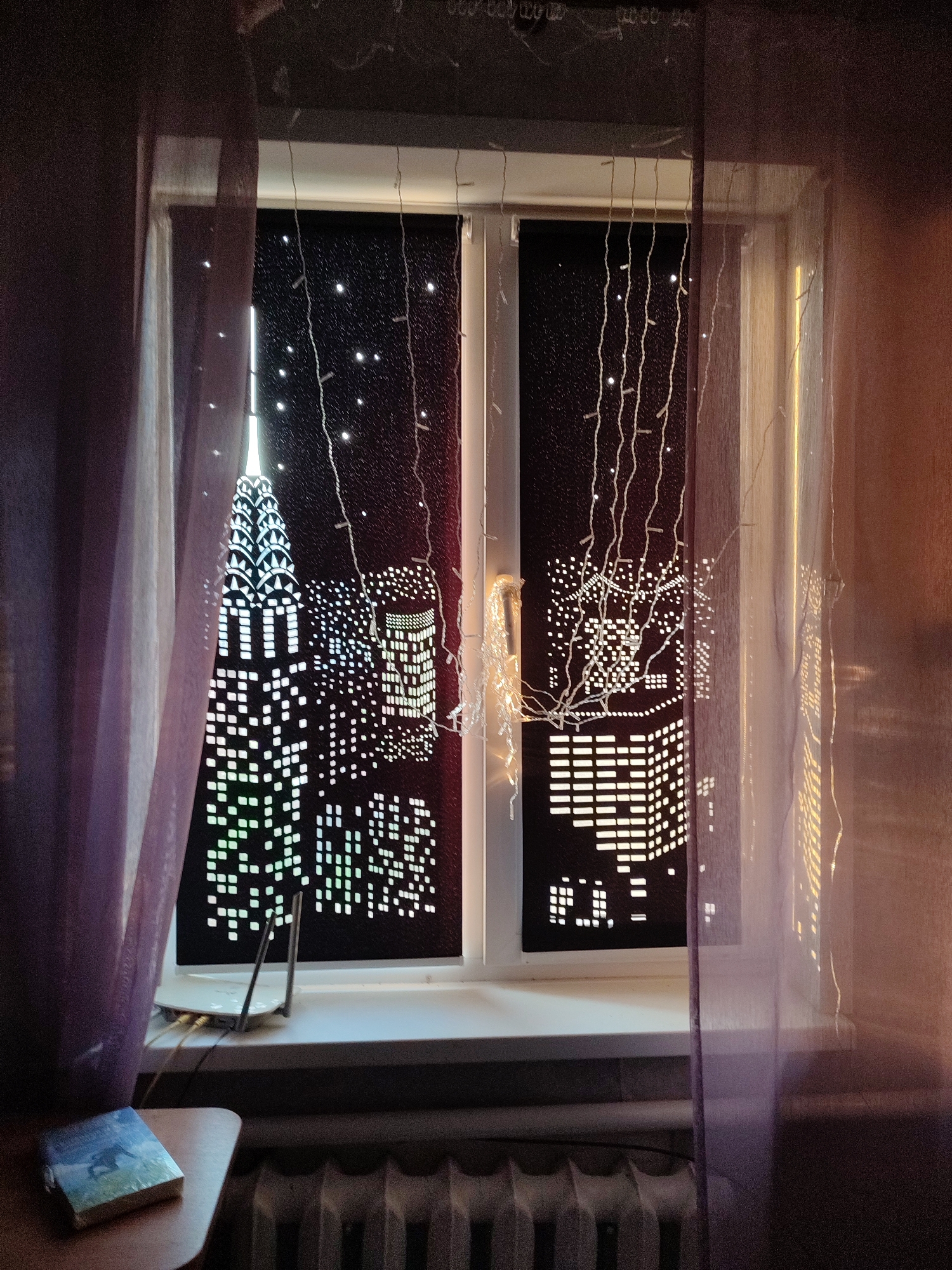 This media file depicts a window with a nighttime cityscape scene created on the blinds. The blinds are dark with cutouts that allow light to shine through, forming the shapes of skyscrapers and city lights. In the foreground, sheer purple curtains hang on either side of the window, partially obscuring the view. String lights are also draped across the window, adding to the nighttime ambiance.
On the windowsill, a white router with two antennas is visible, along with some cables connected to it. To the left of the window, on a small wooden table, rests a book with a blue cover, featuring an image of a person on what appears to be a horse. Below the window, a radiator is visible.
The overall scene conveys a sense of a cozy indoor setting at night, with a decorative, artificial cityscape providing a backdrop. The lighting, with the string lights and illuminated city scene, creates a warm and somewhat magical atmosphere. There is no visible text in the media file. The location context provided is Abinsk, Russia.