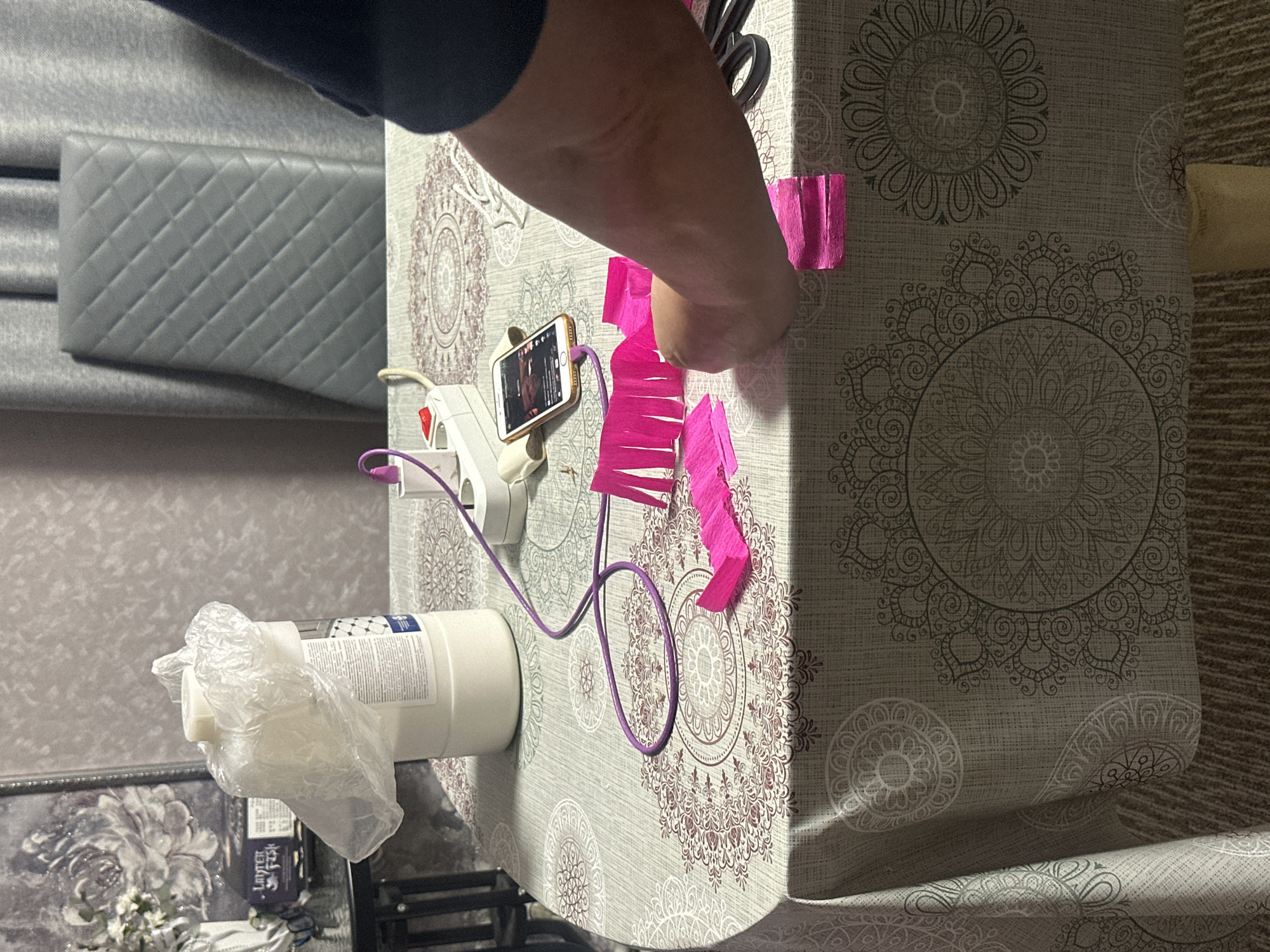 A person is shown on the upper left side of the image using their right hand to handle pink crepe paper on a table with a gray patterned tablecloth in an interior setting. A pair of scissors, a smartphone propped up, a glue gun, and a white plastic-wrapped container with text are also on the table. A gray quilted headboard is visible on the left, and a patterned wall and carpet are in the background. The city and location are unknown.