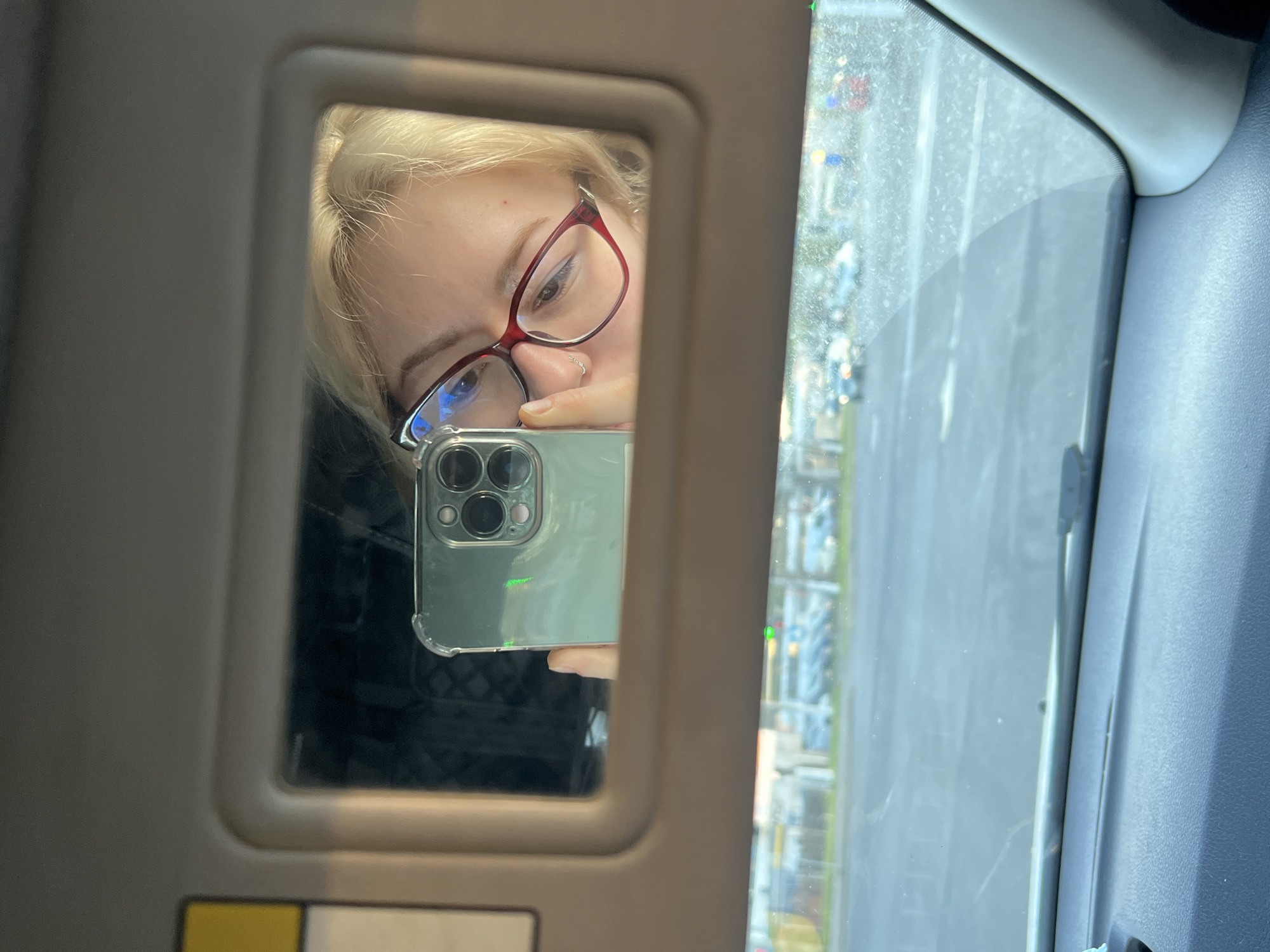 A fair-skinned person with blonde hair and red glasses is taking a selfie in a car mirror. The reflection reveals part of their face, holding a phone with three camera lenses. The background shows a blurry cityscape, possibly Odesa, Ukraine, seen through the car window. The car's interior is visible, with a gray dashboard and sun visor.