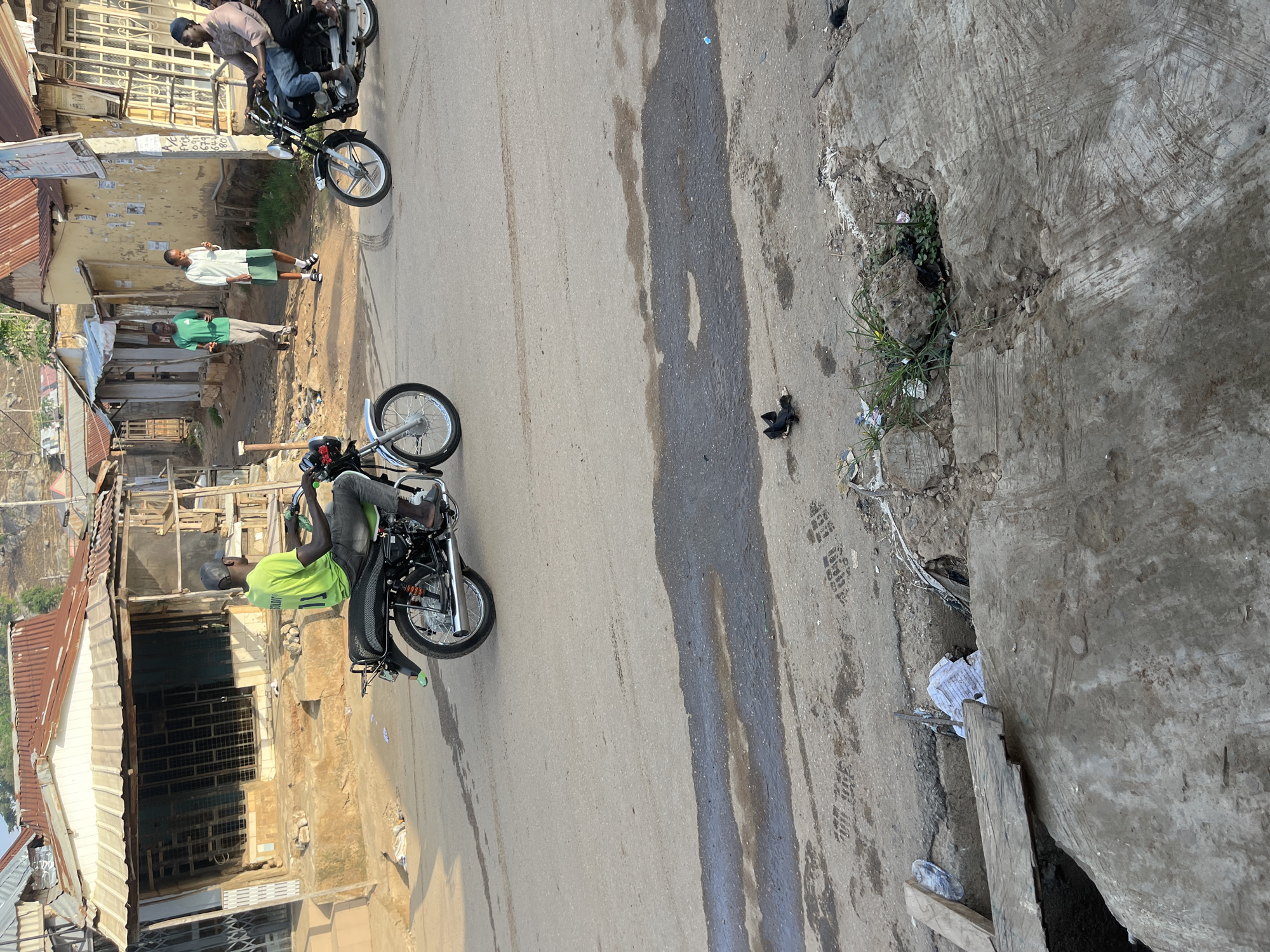 In an urban setting in Abuja, Nigeria, an elderly man wearing a green t-shirt and a grey baseball cap is riding a motorcycle. The motorcycle is black with chrome accents and has a license plate visible. The man is looking ahead as he rides. Behind him, two people are walking. One is a man wearing a green t-shirt and dark pants, while the other is a woman wearing a white shirt and a green skirt, holding something in her hand. Further in the background, another motorcycle is parked, with two people on it. The street is paved and there are buildings on either side, with corrugated iron roofs. Some of the buildings appear to be shops. The area shows signs of wear and tear, with debris on the street and some of the buildings in disrepair.